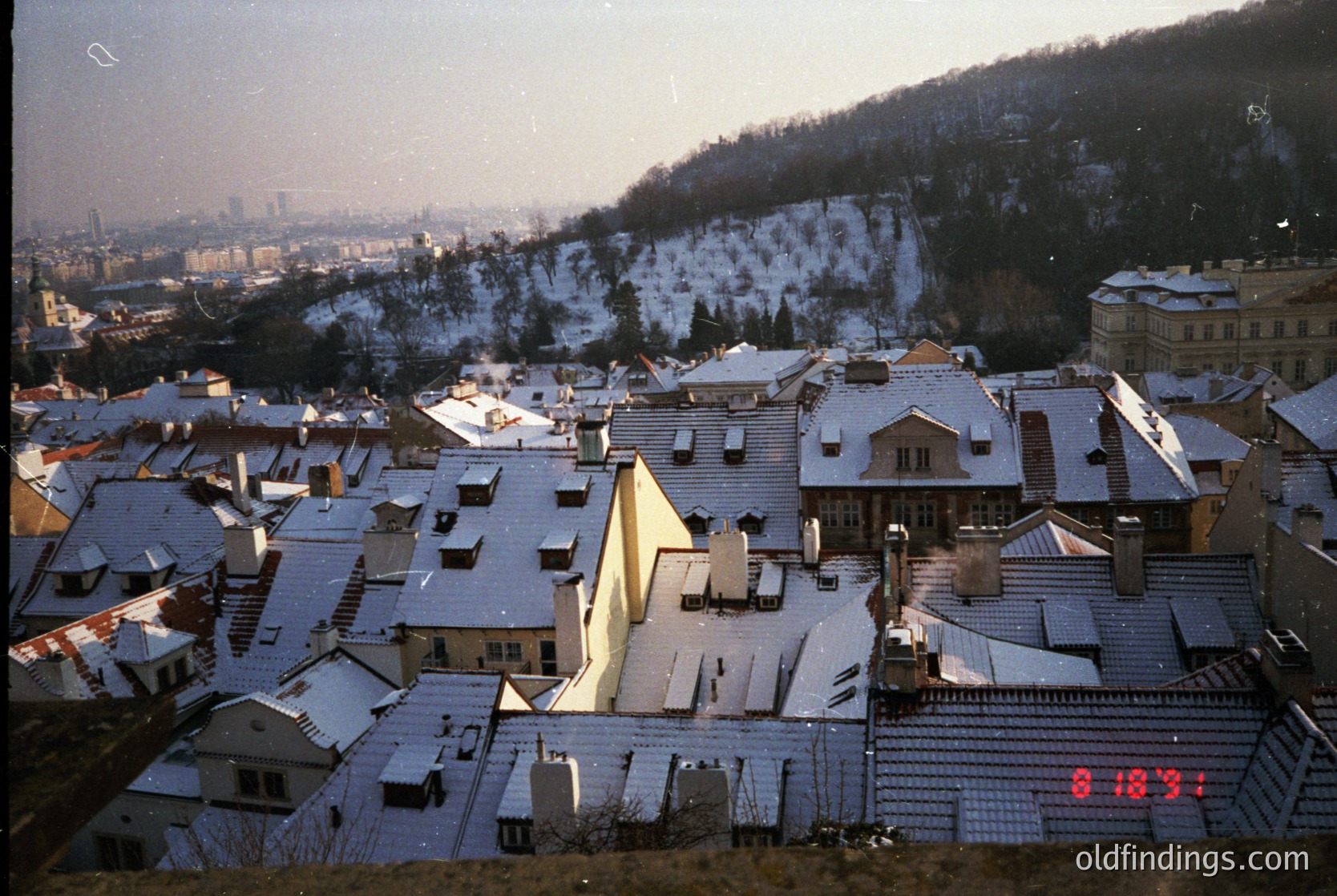 Snow-covered rooftops of a European hillside town, framed by forested hills and distant city skyline. Mid-20th century architecture with sloped roofs and chimneys. Overlay timestamp "8-1973" suggests late 1970s winter scene.