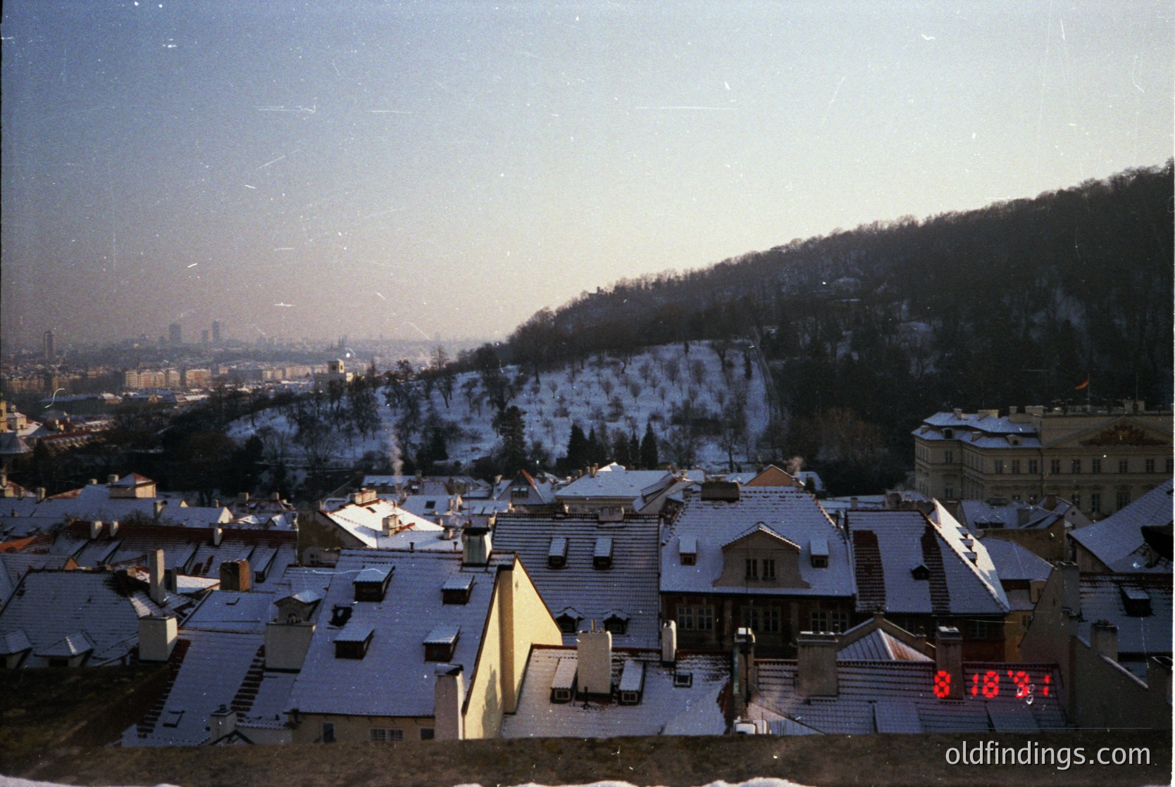 Vintage aerial view of a snow-covered European cityscape, likely . Residential rooftops and terraced hillsides frame a distant industrial skyline. Overlaid digital clock reads 08:18.
