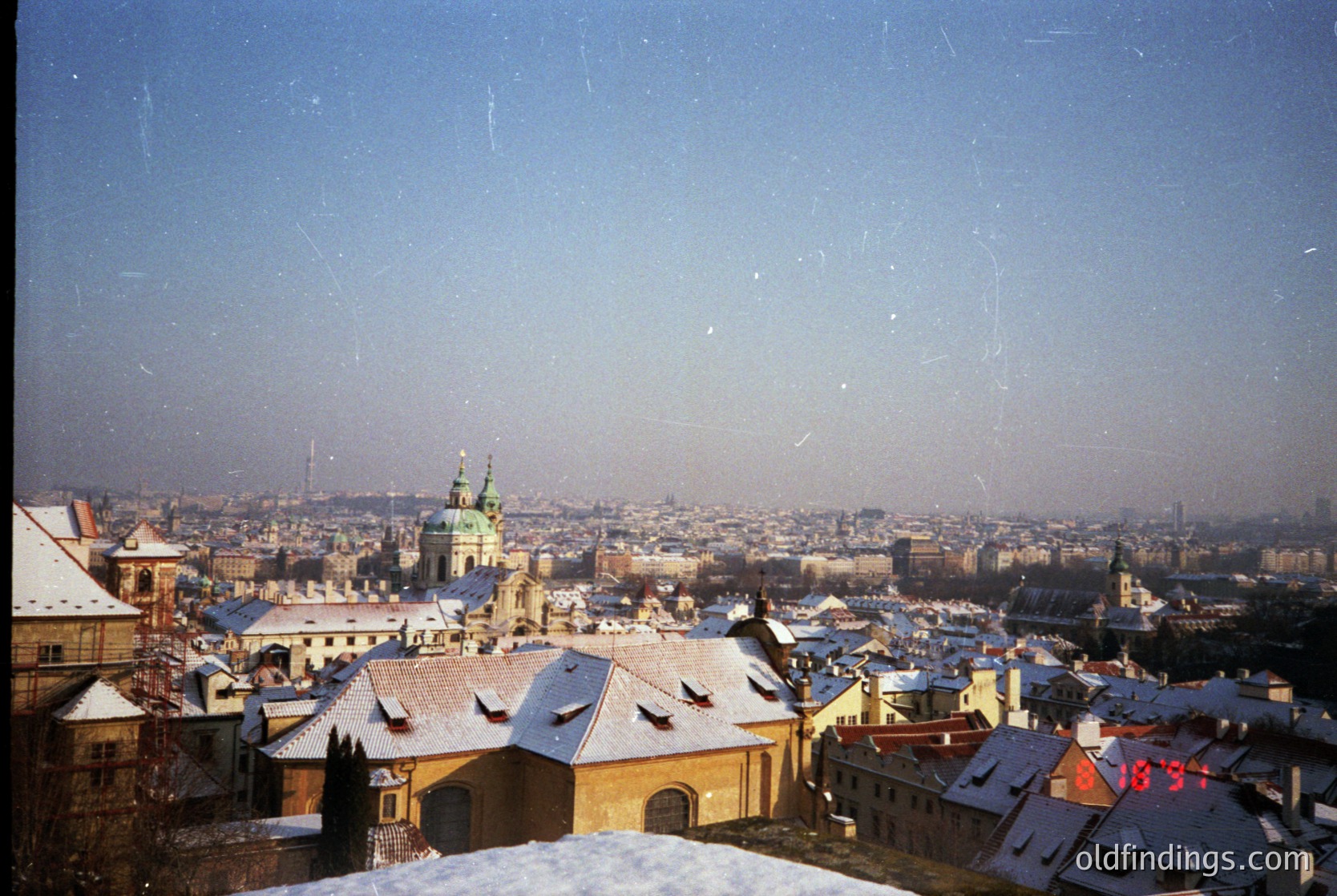 Vintage panoramic shot of Prague’s historic rooftops dusted with snow, showcasing Baroque architecture. Prominent spire and domes hint at Prague Castle’s influence. Urban landscape stretches under clear skies, suggesting early 20th-century or late 19th-century framing.