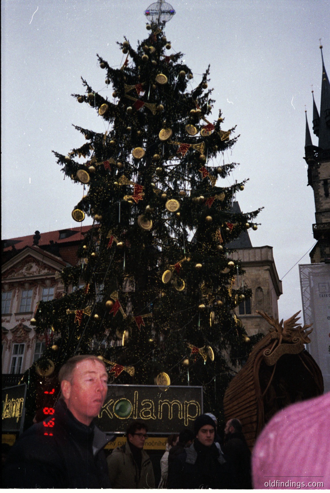 Vintage Christmas scene featuring a towering, elaborately decorated pine tree adorned with golden ornaments and a large reindeer figure. A man holds a sign reading "Kolamp" in front of a historic European building with ornate architecture. Crowd and festive lights suggest a 1960s-1970s holiday market atmosphere.
