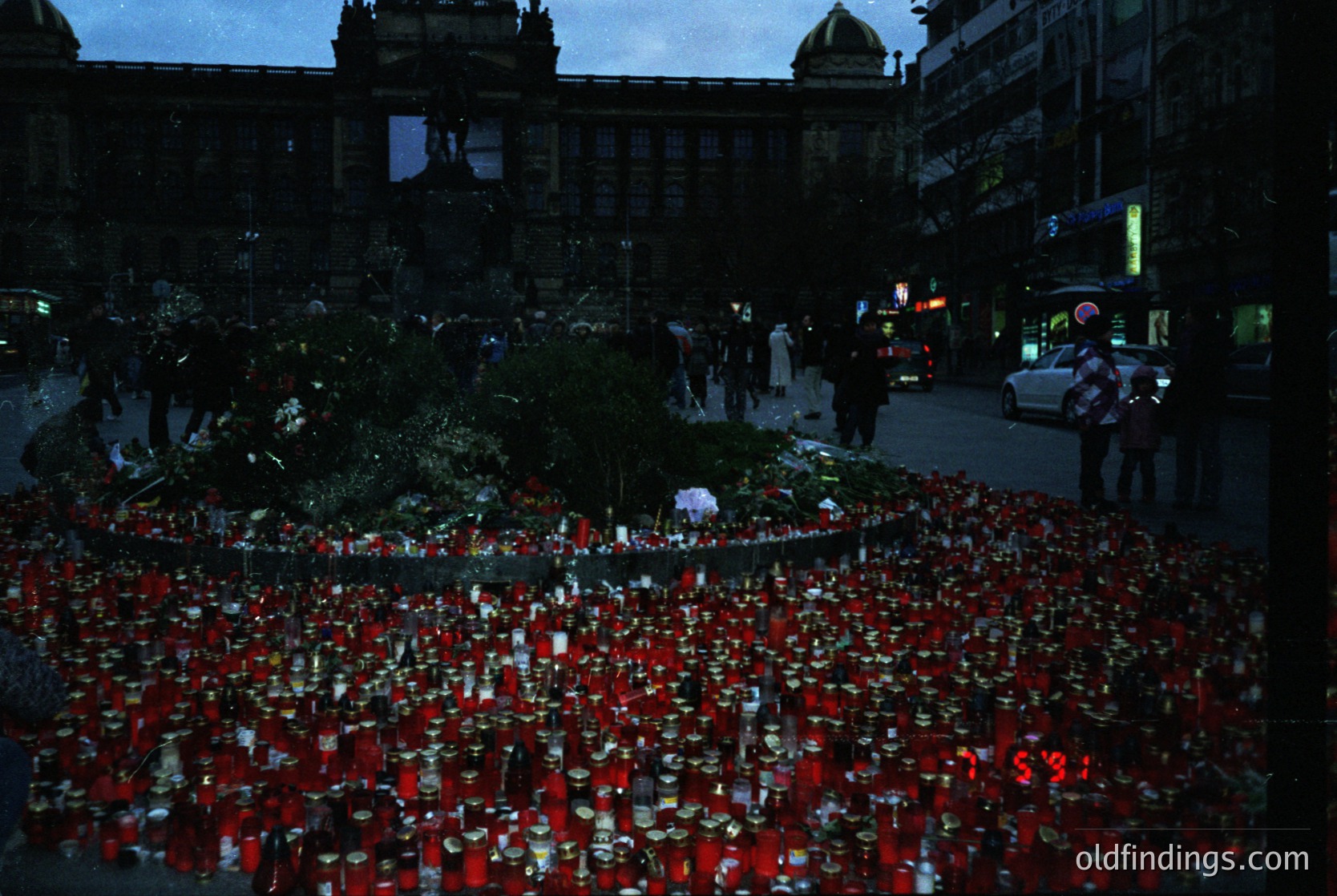 A memorial display of red candles and flowers arranged in a grid pattern on a city street at night, likely honoring a tragic event. Historic European architecture with domed roofs and ornate facades in background. Crowds of people gathered, some holding candles. Urban setting with visible streetlights and parked cars.