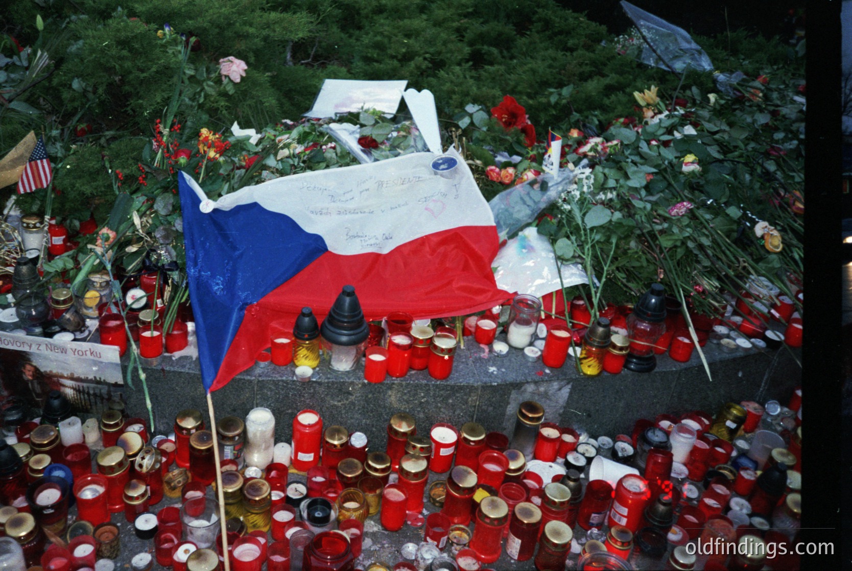 Memorial tribute featuring Czech flag, candles, and floral arrangements. Likely post-2016 terrorist attack in Prague. Crowd-sourced memorial with handwritten notes, international flags, and candles in red cups. Symbolizes solidarity and remembrance.