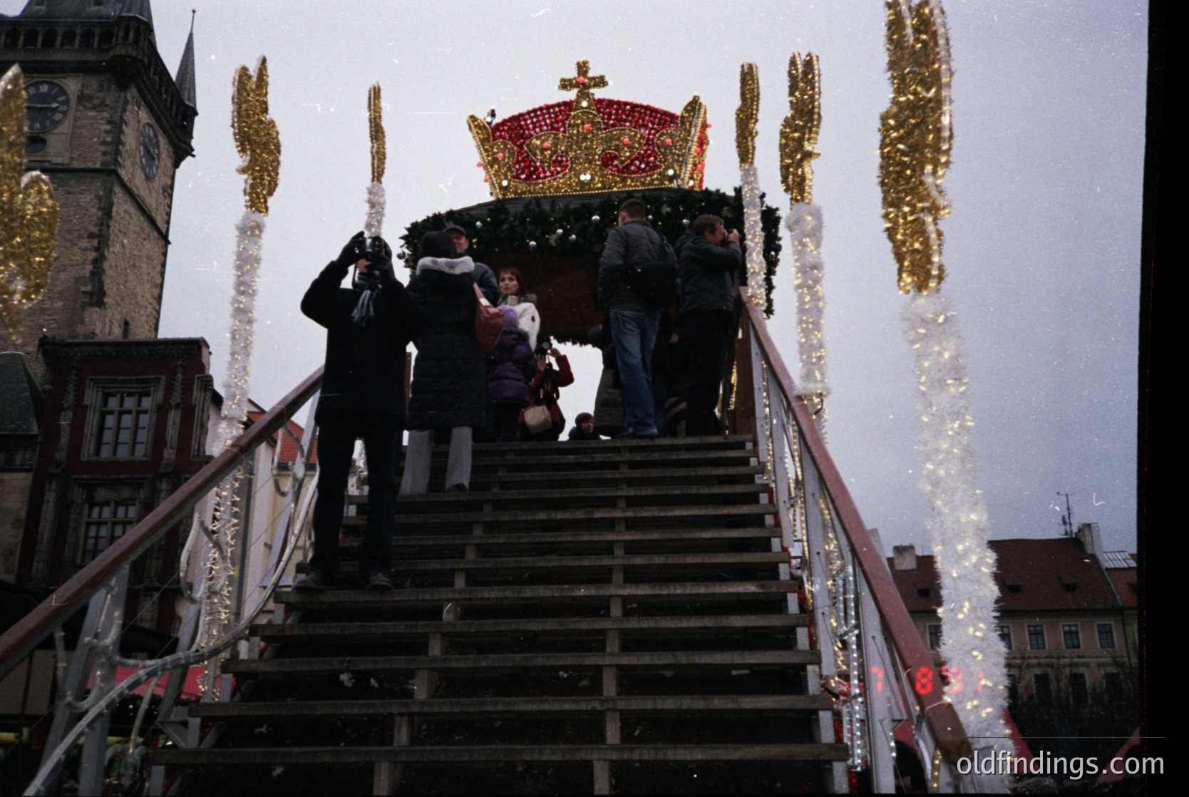 A festive procession ascends a wooden staircase toward a grand, illuminated throne-like structure adorned with a golden crown and red velvet draping. Decorative garlands with golden ornaments flank the stairs. The background features historic European architecture with a prominent clock tower. Crowd wears winter attire; time period appears to be modern (21st century). Likely a holiday or cultural festival.