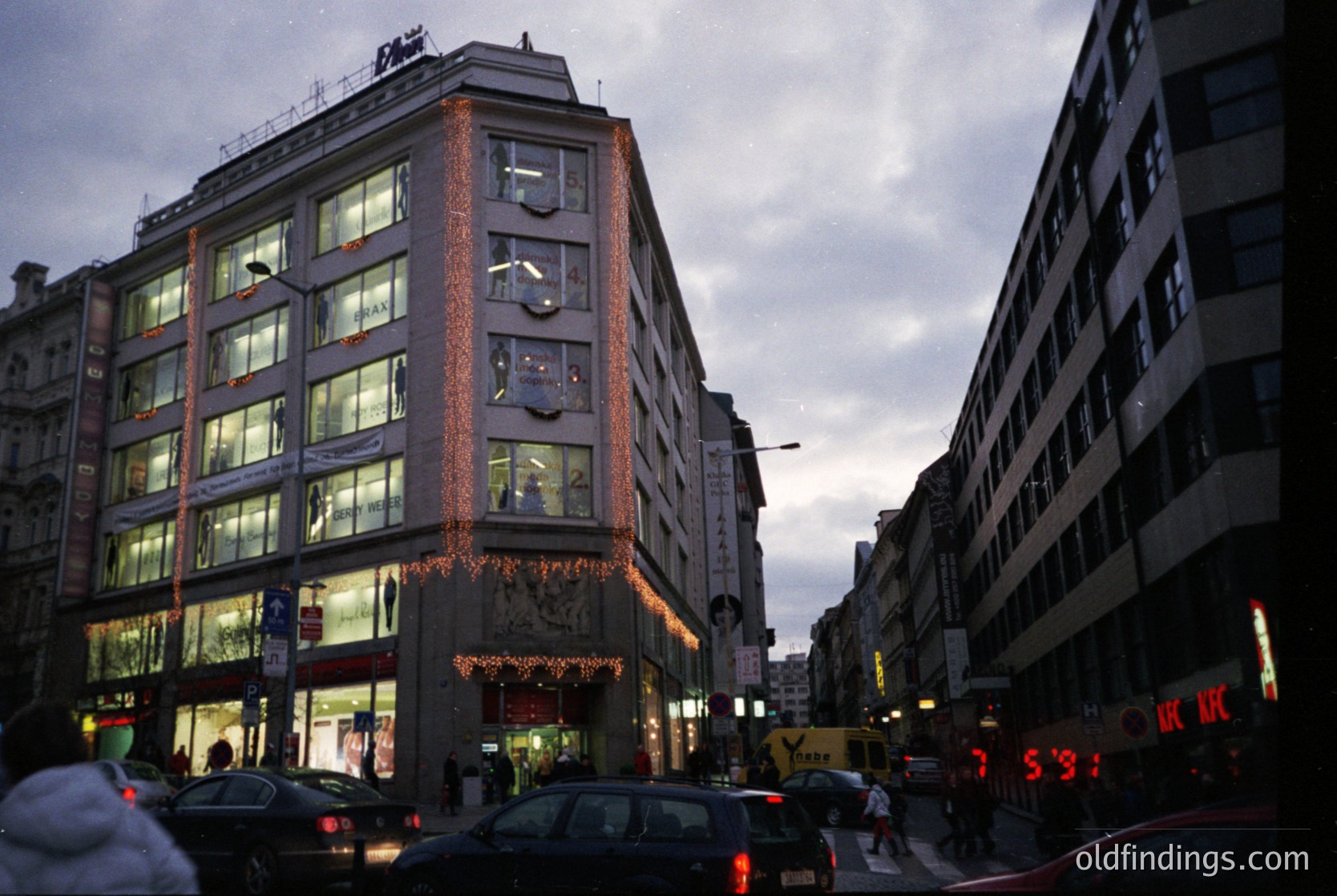 Mid-20th century urban street scene featuring a corner building with illuminated signage and festive garlands. Brick facade with large windows showcases retail or office spaces. Neon signs ($3) and vintage cars suggest a European cityscape, likely 1960s–1970s. Overcast sky and street activity highlight bustling city life.