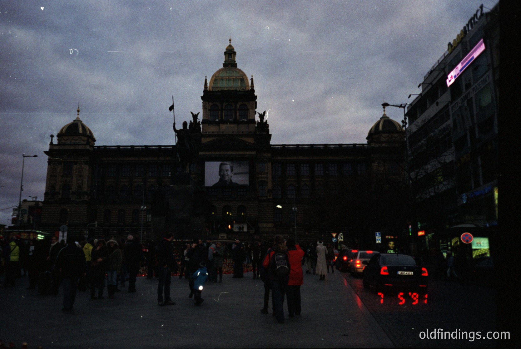 Neoclassical building façade with domed central tower, flanked by smaller domes, illuminated by large portrait projection. Urban street scene at dusk, featuring pedestrians and traffic. Likely a public square or central plaza.