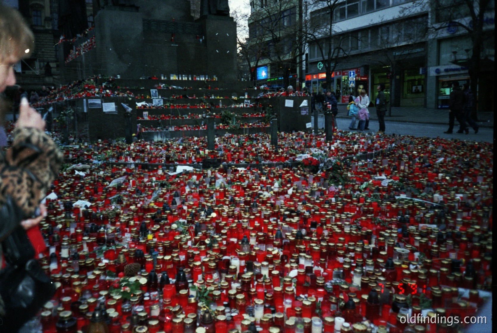 Memorial tribute featuring thousands of red candles and candles in jars arranged in a dense, organized pattern on a city street. Urban setting with multi-story buildings and streetlights in background. Likely commemorative event, possibly post-disaster or tragic incident.
