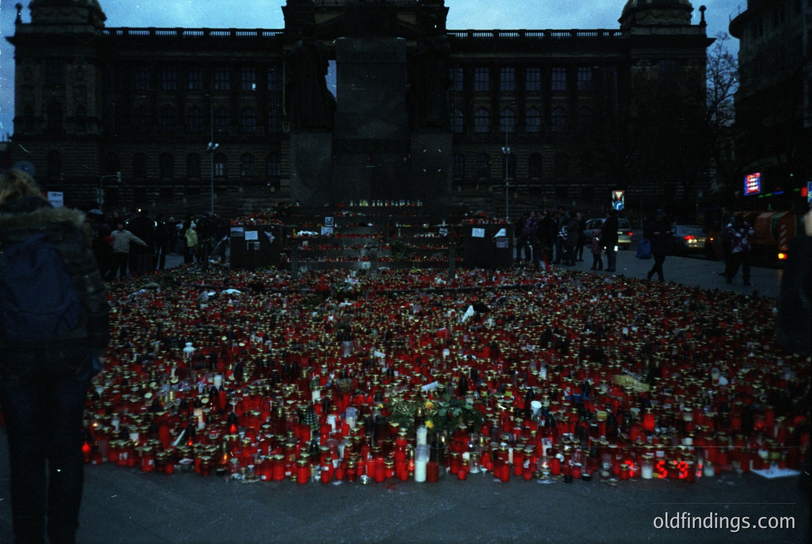 Memorial tribute featuring thousands of red roses and candles arranged in a dense, heart-shaped pattern on a city plaza. Surrounding historic building with ornate architecture in the background. Likely a public vigil or remembrance event.
