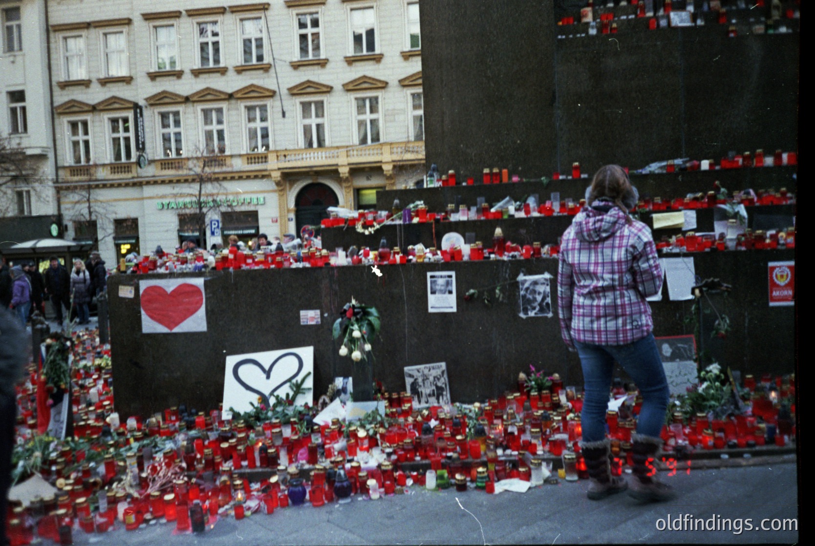 Memorial tribute wall lined with red candles, flowers, and handwritten notes in an urban plaza. A large red heart symbol dominates the center. Architectural backdrop features classic European facades with arched windows. Crowd of people observing from a distance. Likely post-2015 terrorist event in Brussels ( ).