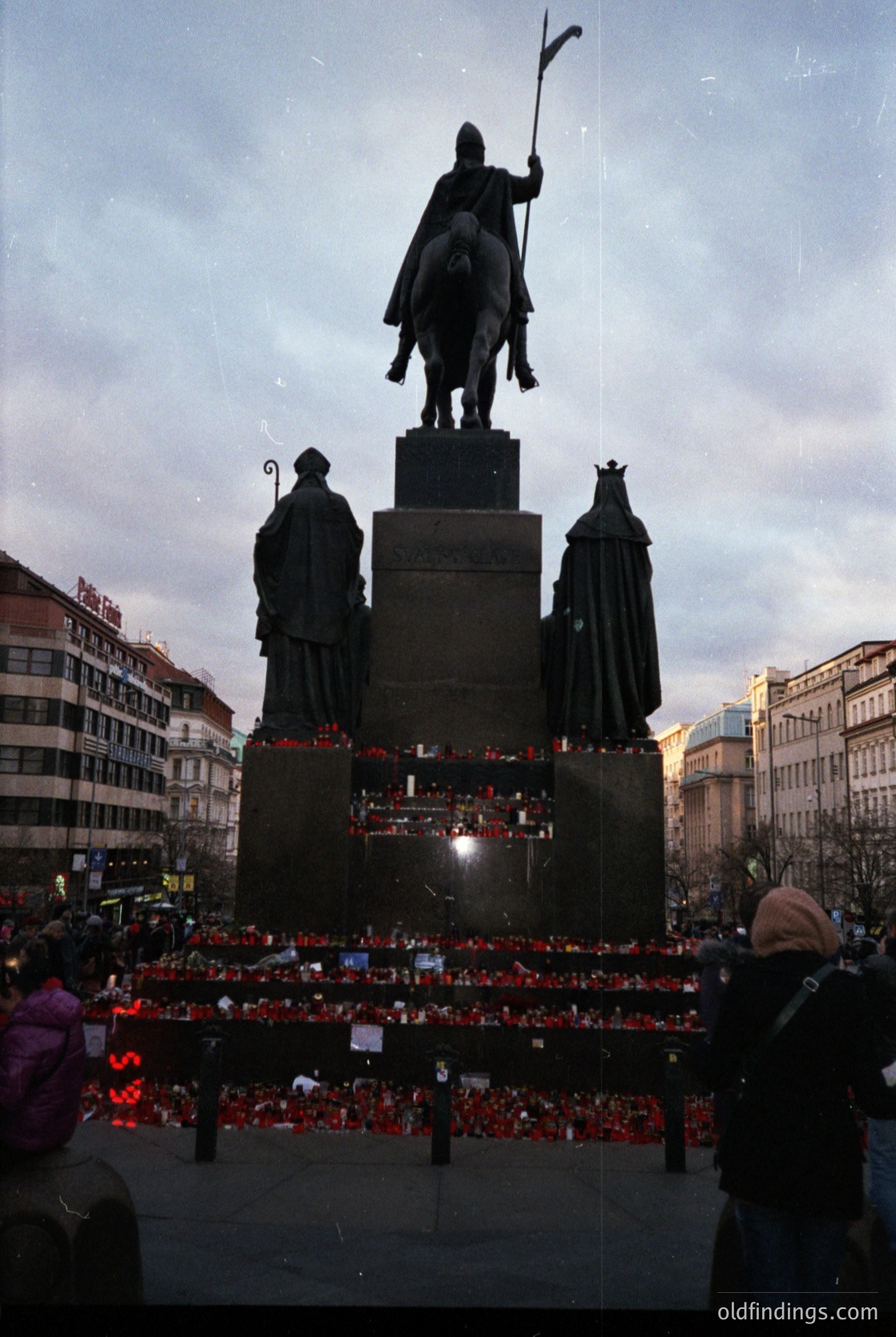 Equestrian statue monument with flanking figures, surrounded by red candles and floral tributes. Urban setting with historic architecture in background. Likely Prague’s Wenceslas Square,