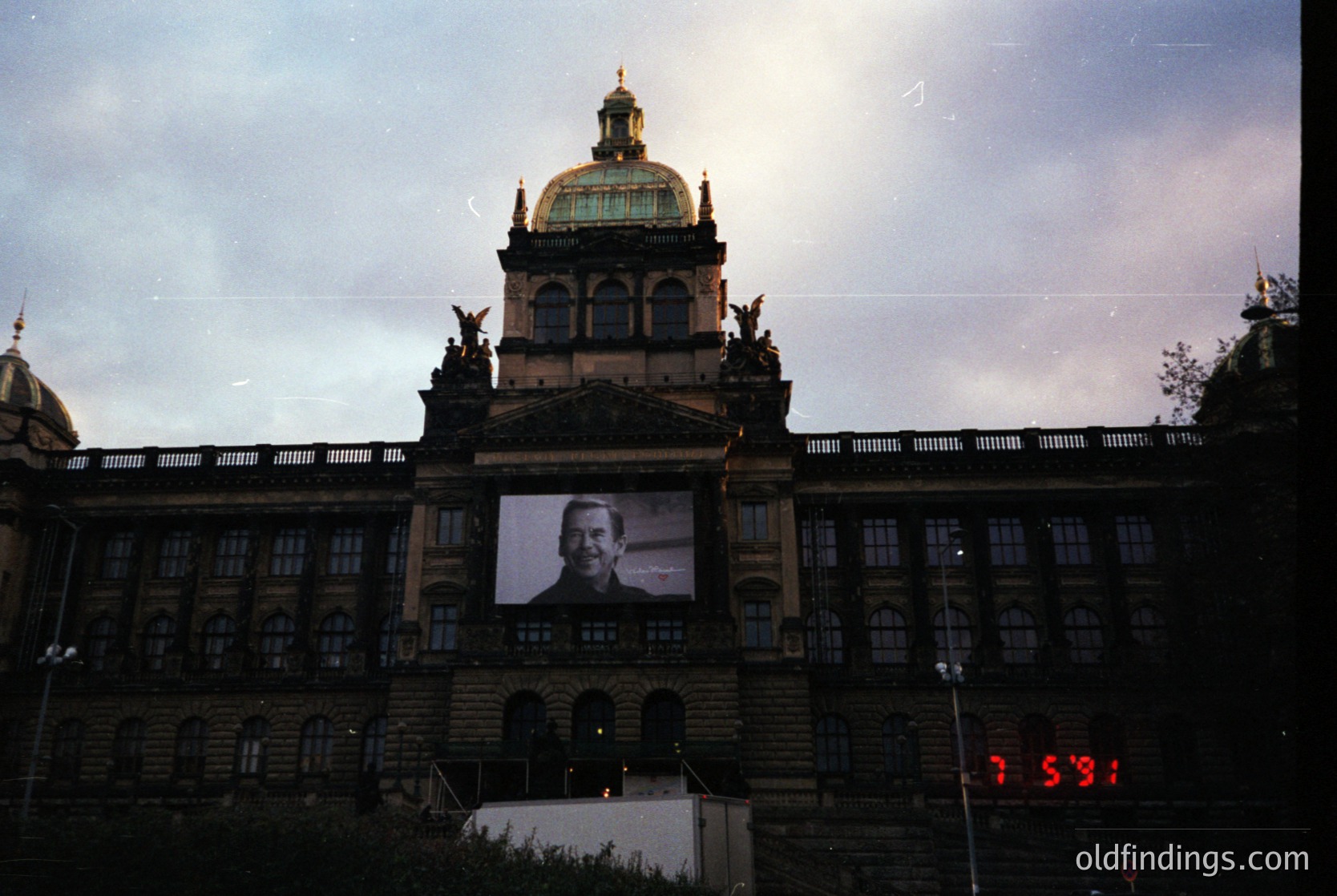 Neoclassical building with domed roof and statues, featuring a large projected portrait of a man on its facade. Likely Prague’s National Theatre, 1960s–70s era. Dramatic evening lighting enhances architectural details.
