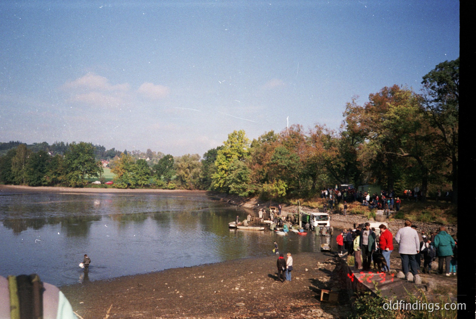 Vintage lakeside gathering with dense foliage framing a calm water body. Group of people in casual 1970s–80s attire—some wading, others standing near parked vehicles. Wooden dock extends into water, hinting at recreational use. Overcast sky with soft lighting suggests early autumn.