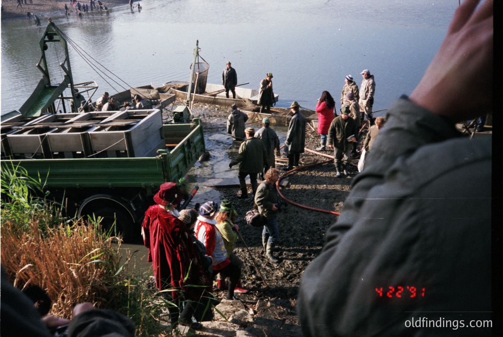 Vintage fishing scene with group unloading nets from a green truck near a waterfront. Mid-20th century attire and equipment suggest rural coastal labor. Overcast skies and natural lighting indicate early/late season.
