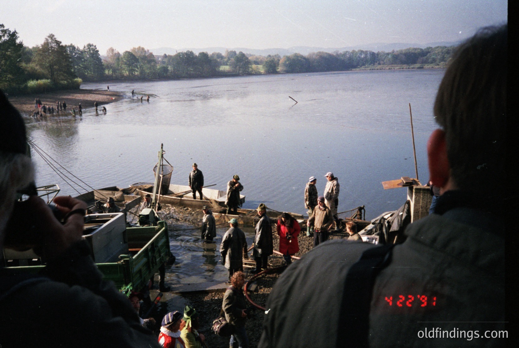 Vintage fishing scene with wooden docks and boats on a calm lake, likely Eastern Europe . Crowd in layered clothing—some holding nets—suggests autumn or early spring. Industrial-era fishing equipment visible.
