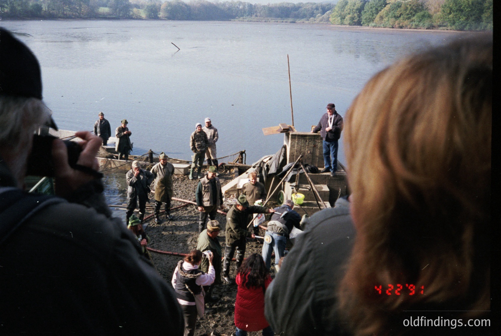 Rustic lakeside scene featuring a wooden dock with a makeshift fishing platform. Crowd of onlookers, including children, gathered on shore. Adults in casual attire—some in jackets—engage in conversation or observe. Overcast sky reflects on calm waters. Timestamp "12:33" visible in bottom-right corner.
