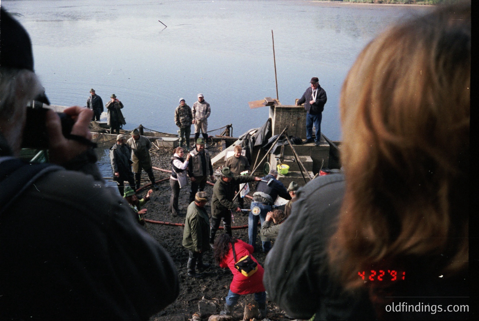 A group of people on a wooden pier by a lake, likely for a ceremonial or public event. Individuals wear 1970s-era clothing (oversized jackets, vests, and headscarves). One person in a red jacket kneels, possibly filming or documenting the scene. The pier’s structure and crowd suggest a formal gathering, possibly tied to local traditions or government activities.