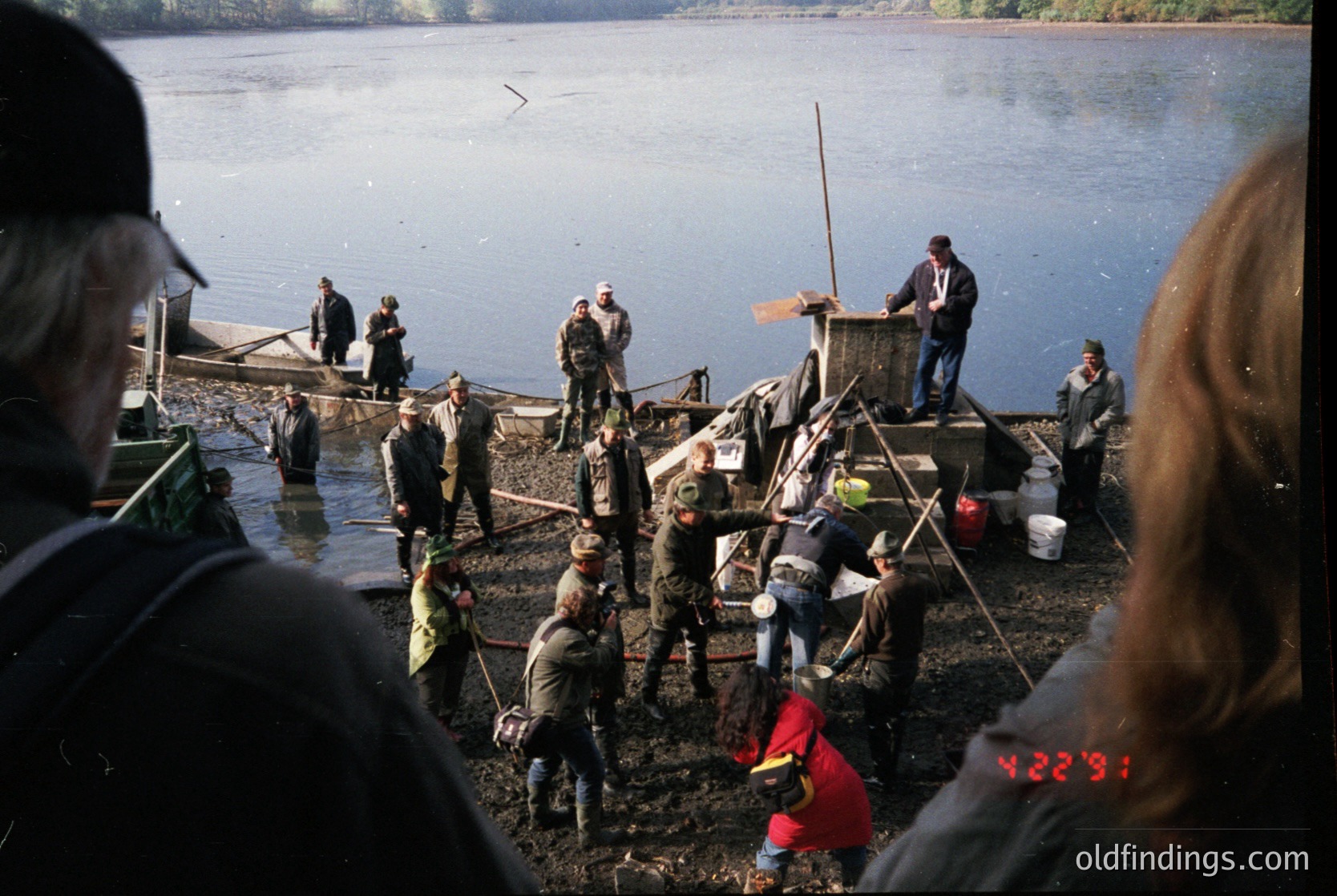 Vintage black-and-white photo of a communal fishing operation on a muddy shore. Men in workwear (overalls, caps) and a few women assist with hauling nets from a small wooden dock into shallow water. A makeshift pulley system and buckets suggest manual labor. Background shows a calm lake and forested hills. Likely Eastern Europe, 1960s–1980s.