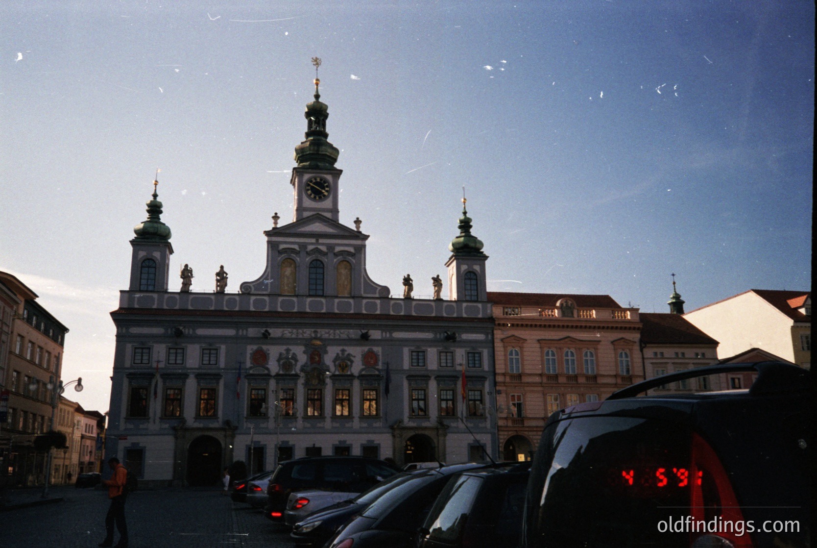 Baroque-style town hall with ornate façade, featuring a central clock tower and sculptural details. Symmetrical windows and decorative reliefs dominate the upper levels. Parked cars and modern vehicles frame the scene, suggesting urban setting. Likely European, possibly Central/Eastern. #