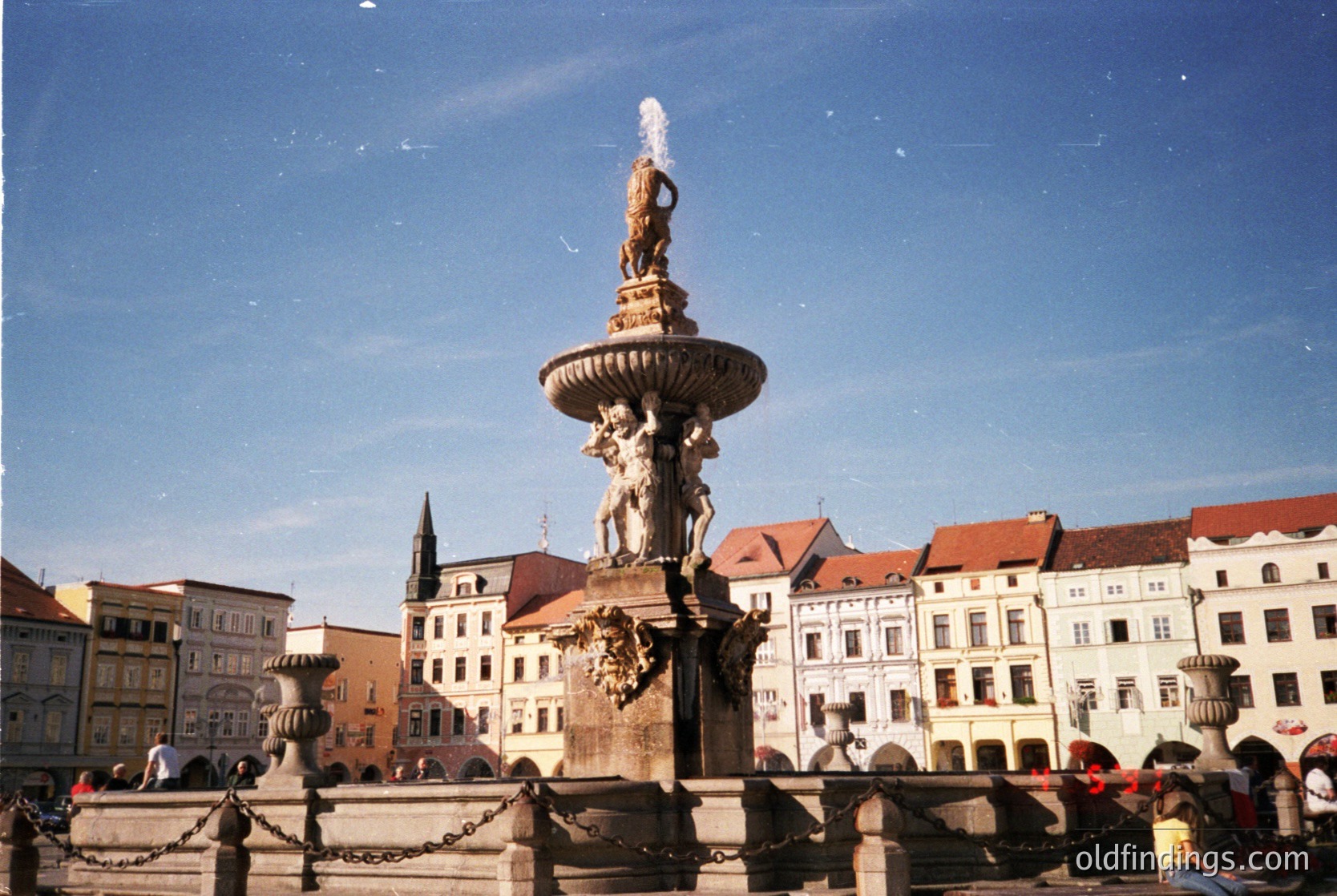 Neoclassical fountain with central statue and cascading water in a European city square, surrounded by multi-story, pastel-colored buildings with red-tiled roofs. Likely European architecture.