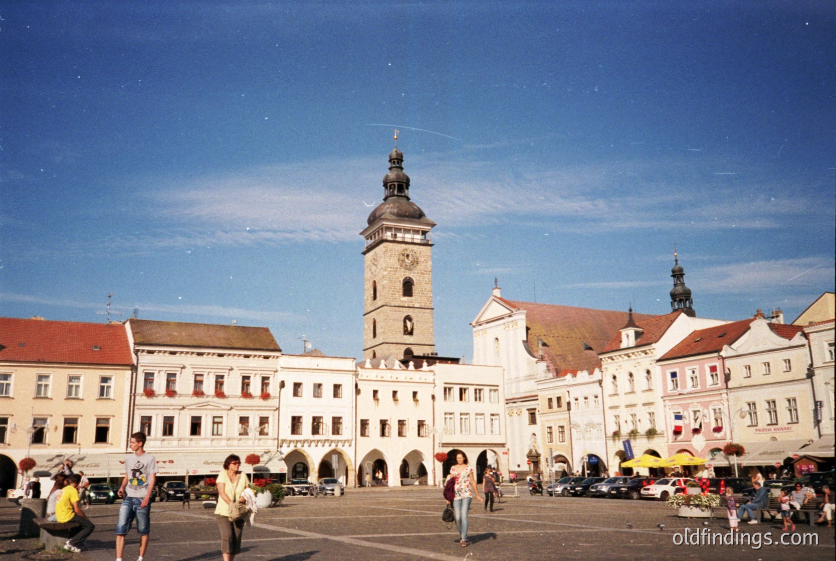 Historic European town square with Baroque-style architecture. Prominent clock tower with spire dominates center, flanked by white facades with red-tiled roofs. Outdoor market stalls and pedestrians populate the plaza under clear skies. Likely