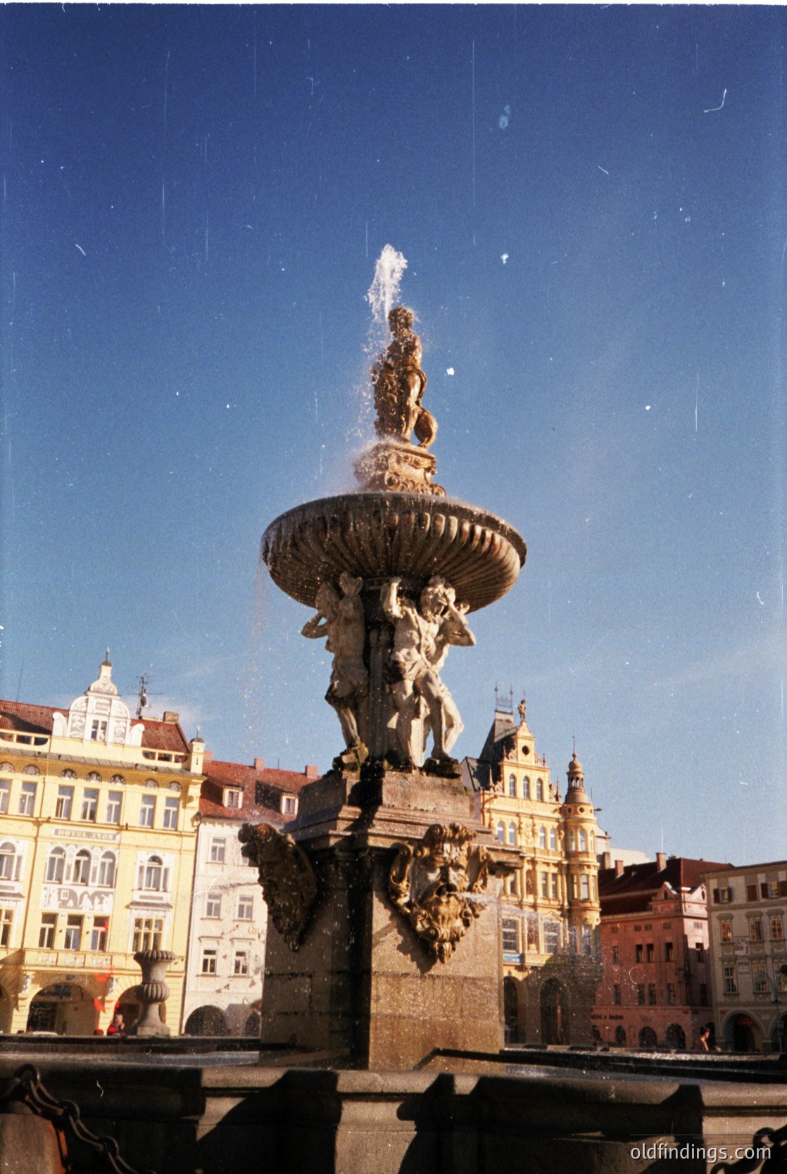 Baroque-style fountain with water spouting from a central urn flanked by sculpted figures, set against historic European architecture. Likely 18th-century design, featuring intricate stonework and classical motifs.