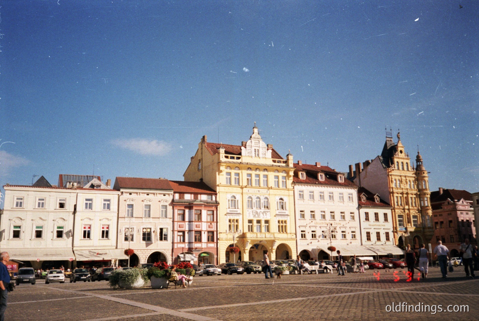 Historic European town square with **Art Nouveau and Baroque** facades. Central building features arched ground-floor entrances and decorative cornices. Cobblestone plaza with outdoor seating, parked cars, and pedestrians. Likely **Czech Republic** (Prague-style architecture).