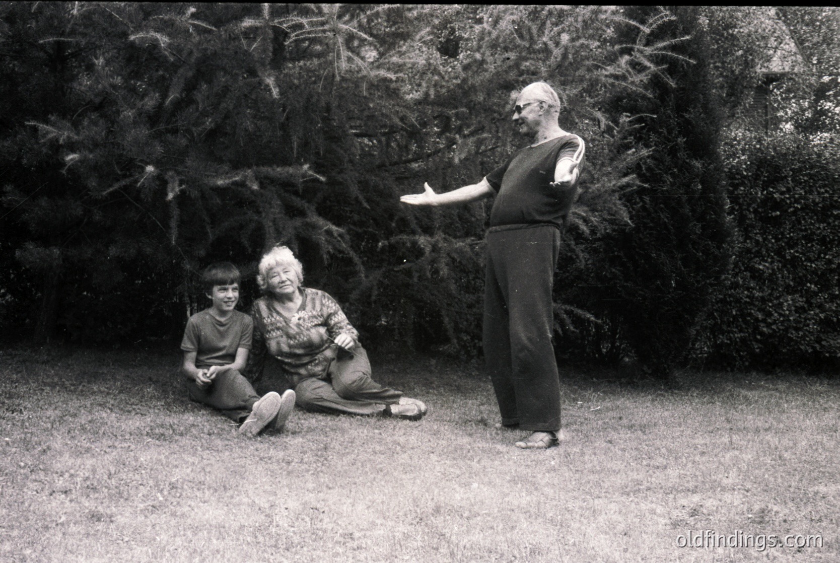 Mid-20th century black-and-white portrait: three individuals in a lush garden setting. A man in a short-sleeve shirt stands gesturing, while a woman in patterned clothing and a child sit on grass beside a dense shrub. Mid-century fashion and outdoor photography style suggest 1950s–1960s era.