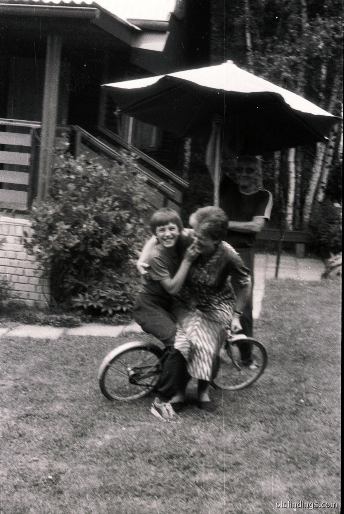 Two children joyfully embrace on a vintage tandem bicycle in a mid-century backyard. The adult in the background under a patio umbrella suggests a casual, family-oriented setting. Likely 1950s–1960s USA.