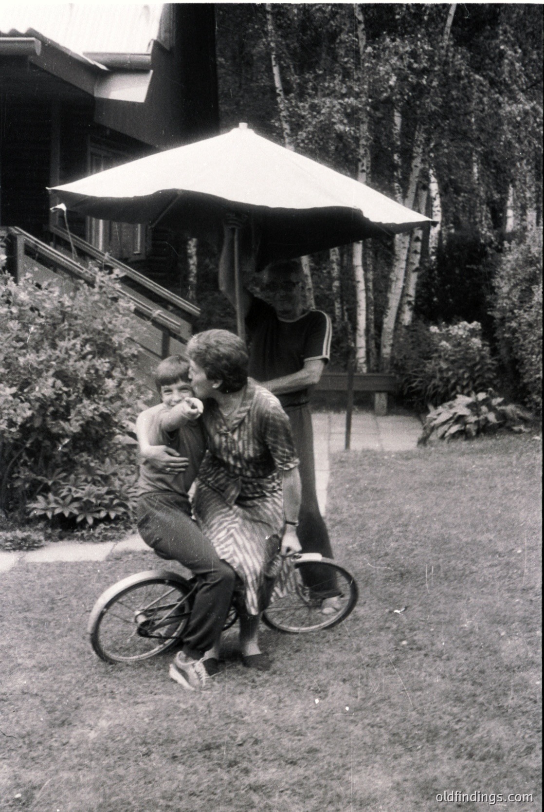 Vintage black-and-white photo of two adults and a child in a lush garden setting, likely mid-20th century. A woman in a striped dress and patterned blouse holds a young child on a stationary bicycle, while another adult stands nearby under a large umbrella pavilion. Wooden deck and greenery frame the scene, suggesting a suburban or resort-style home.