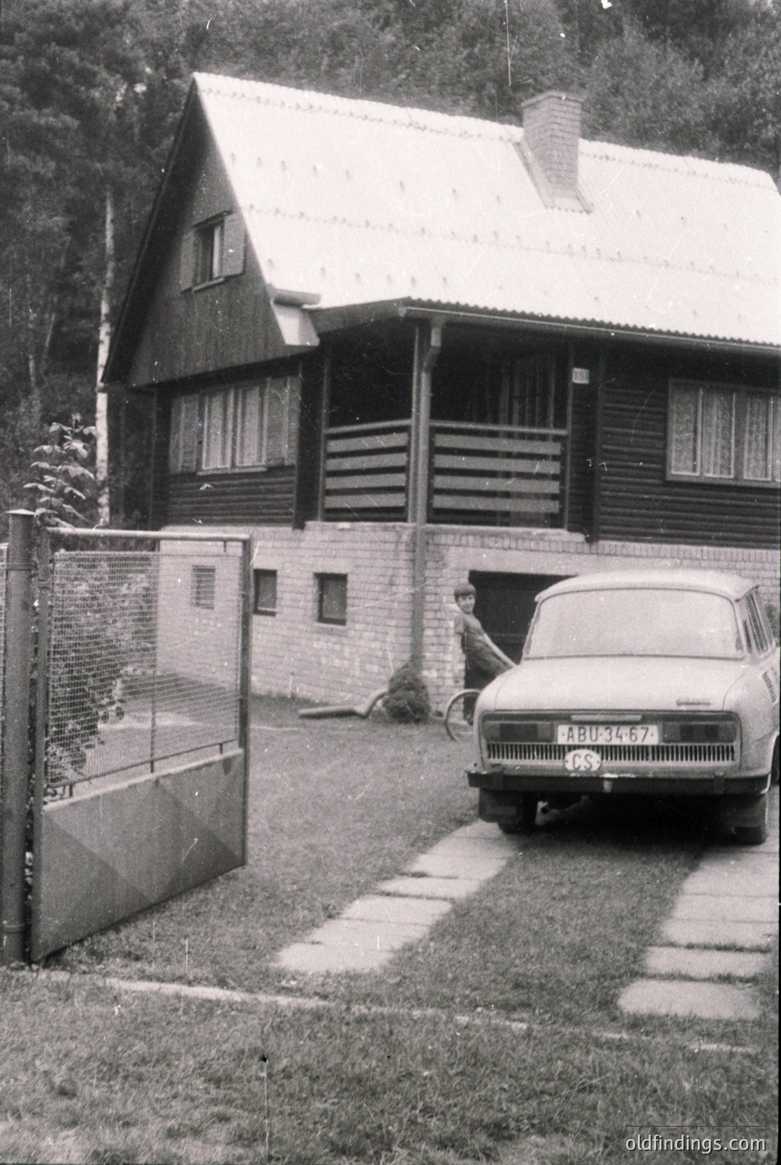 A vintage log cabin-style house with a pitched roof and brick foundation, framed by a simple metal gate. A Soviet-era ZAZ-965 car (1960s-70s) parked beside the gate, with a person standing near the entrance. Lush greenery and trees in the background suggest a rural or suburban setting.