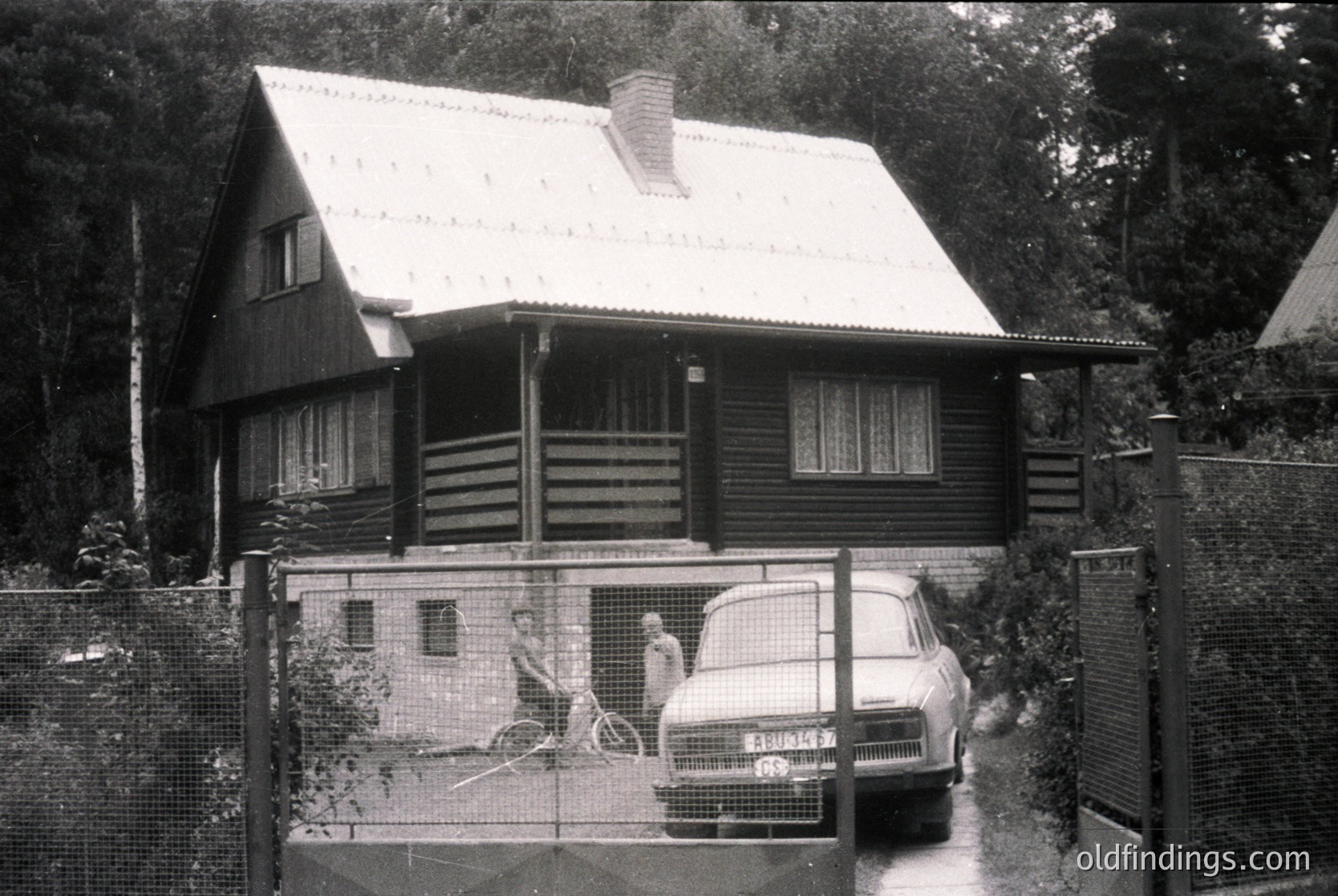 Mid-century wooden house with gabled roof and wooden balcony, framed by chain-link fence. Classic sedan parked beside a child playing on a small elevated platform. Likely Eastern Bloc-era residential architecture, 1960s–1970s.