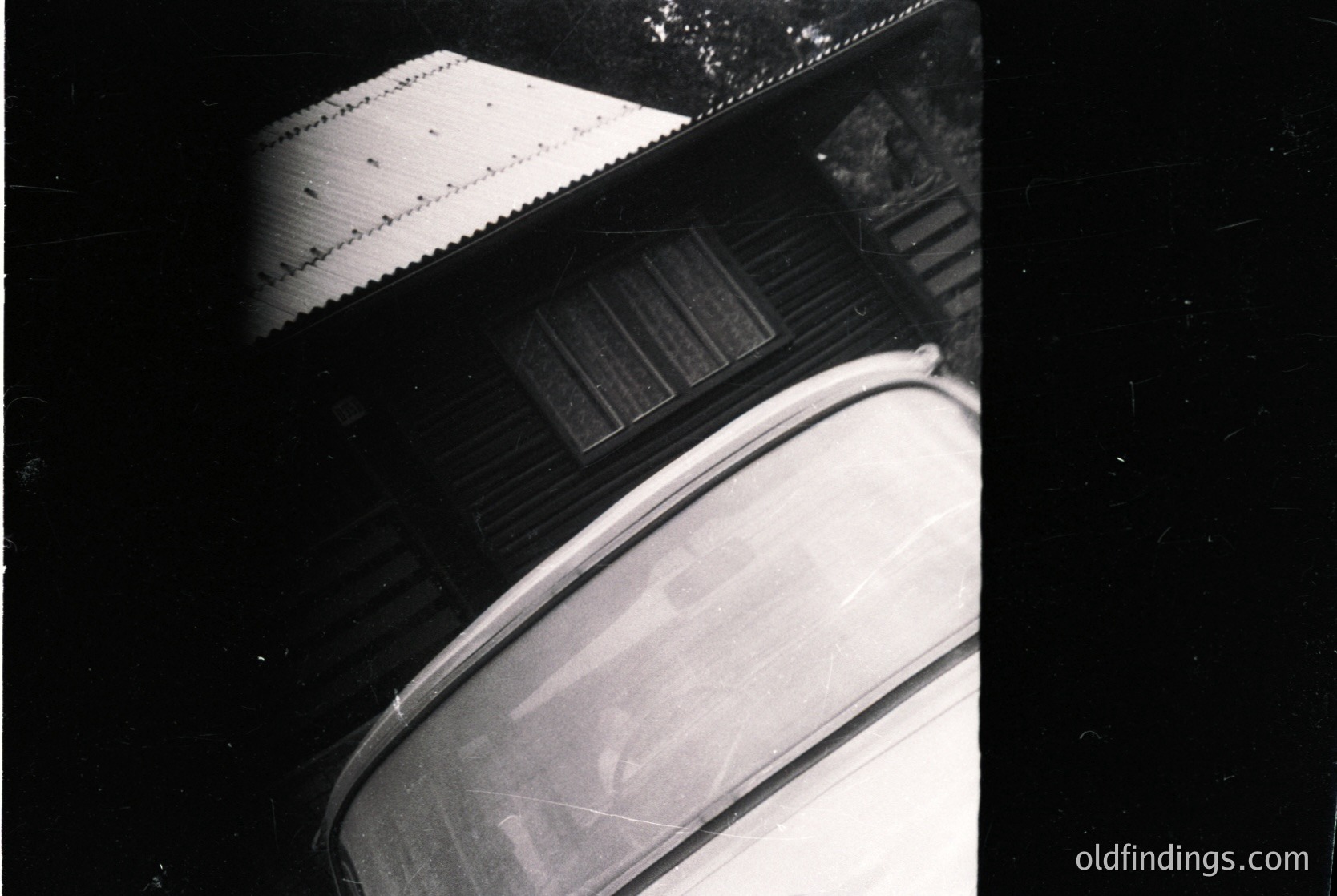 Vintage black-and-white photo of a **1950s-60s** **automobile** (likely a **Volkswagen Beetle**) parked beside a **wooden fence** with **corrugated metal roofing**. Low-angle shot highlights the car’s **rounded rear** and **chrome accents**. Rustic, mid-century industrial aesthetic.