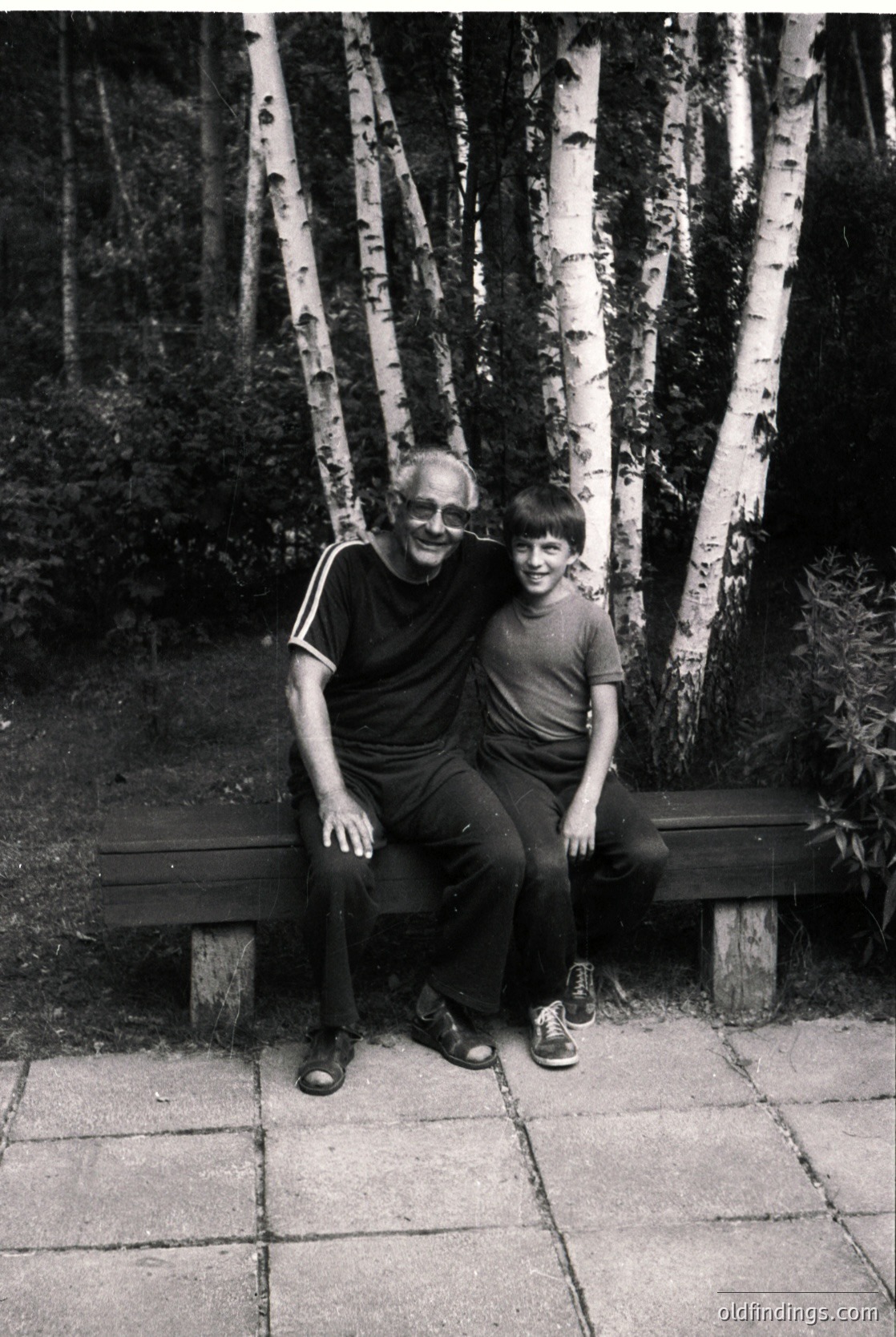 Black-and-white portrait of an adult and child seated on a park bench surrounded by birch trees, likely mid-20th century. The man wears glasses, a striped shirt, and dark pants; the boy sports a short-sleeve shirt and sandals. The paved pathway and natural lighting suggest an urban park setting.