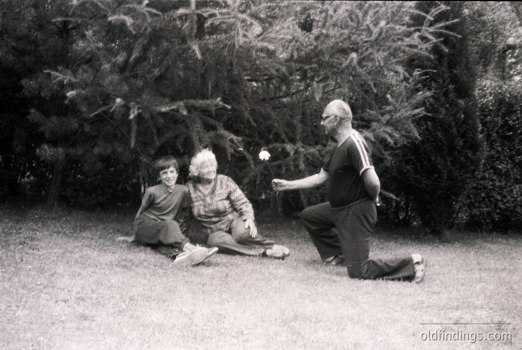 Mid-20th century black-and-white photo: three individuals in a lush garden setting, likely 1950s–1960s. A man kneels, tossing a flower; a woman sits beside him, holding a patterned shawl; a child sits nearby, observing. Dense greenery and grassy lawn frame the scene.