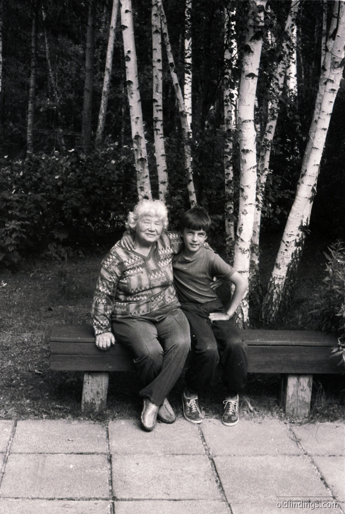 Black-and-white portrait of an elderly woman and young boy sitting closely on a wooden bench in a birch forest. The woman wears a patterned sweater and dark pants; the boy sports a striped shirt and sneakers. The birch trunks create a natural frame. Likely mid-20th century, possibly or .