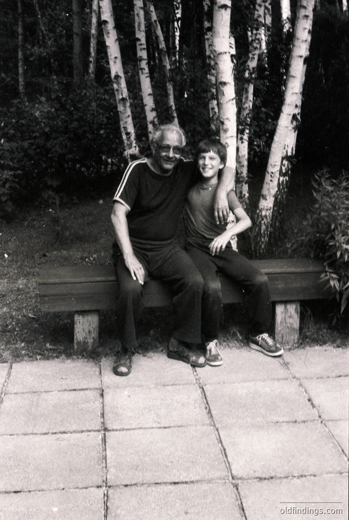 A man and woman pose closely on a wooden bench in a birch forest, likely 1970s–1980s. The man wears a striped polo shirt and glasses; the woman sports a sleeveless top and a necklace. Pathway tiles and birch bark details add texture.