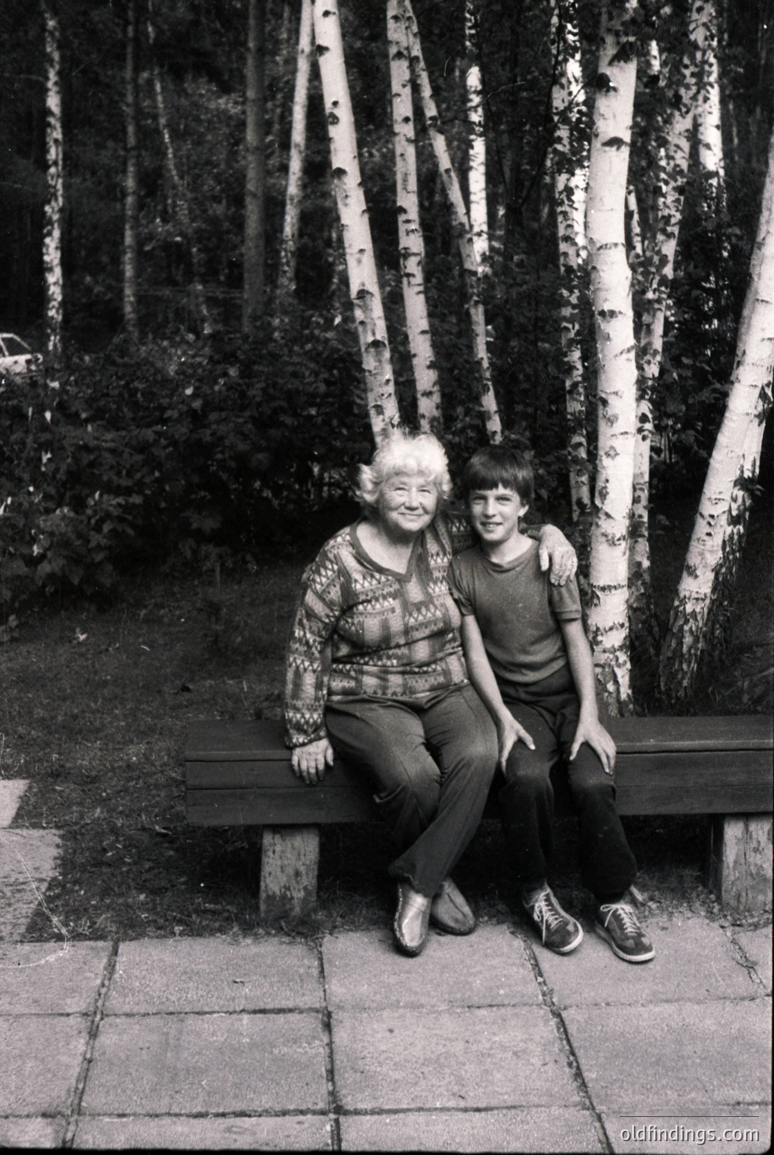 A black-and-white portrait of an elderly woman and young boy seated on a wooden bench in a birch forest. The woman wears a patterned sweater and trousers; the boy sports a striped sweater and sneakers. Birch tree trunks dominate the background, creating a serene, natural setting. Likely mid-20th century, possibly for nostalgic or family history research.
