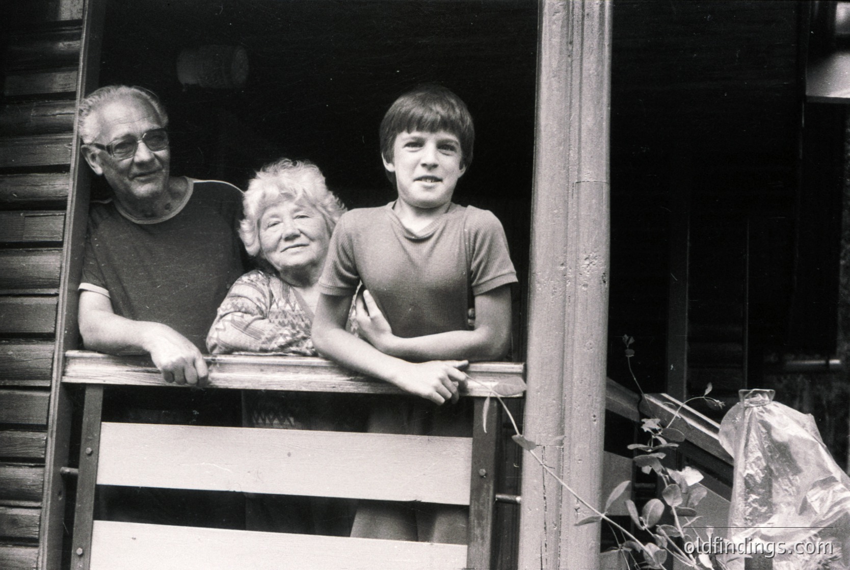 Three generations pose on a wooden balcony, framed by rustic wooden railings. Elderly man and woman in casual 1970s attire (turtleneck, glasses, floral blouse) flank a boy in a short-sleeve shirt. Indoor light casts soft shadows; plastic bag and greenery hint at domestic life.