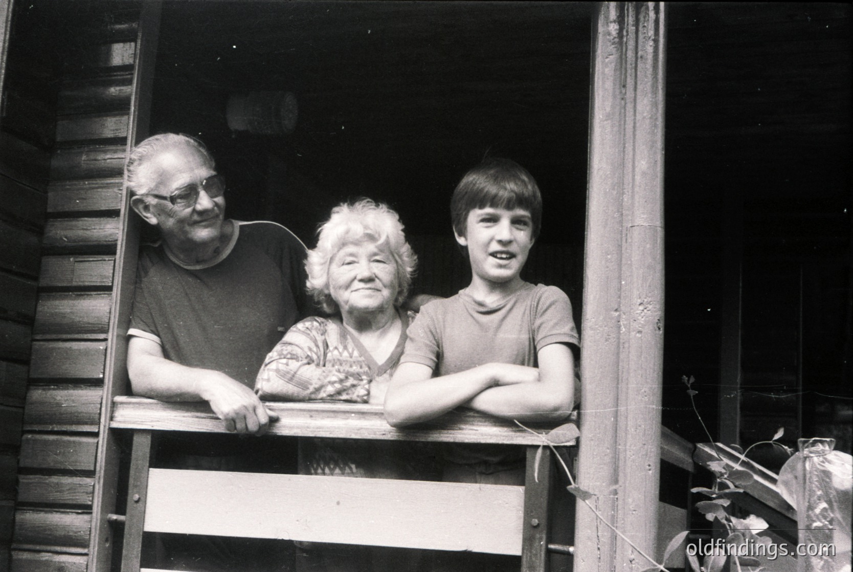 Three-generation family poses on rustic wooden balcony, likely Eastern European rural setting. Elderly man, woman, and young boy in casual 1970s attire. Wooden planks and simple construction suggest modest living. Warm, intimate moment captured in black-and-white.