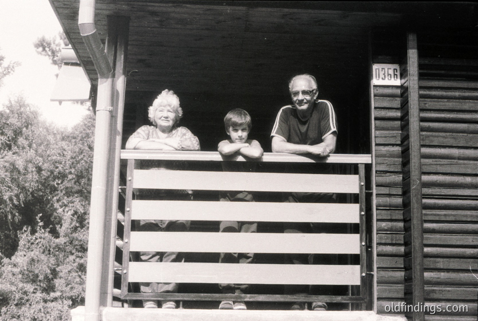 Family portrait on a wooden porch, likely mid-20th century. Three generations pose on a wooden balcony with a rustic wooden house (, ). The man’s striped shirt and woman’s patterned sweater suggest casual, everyday attire. House number "135" visible on siding. Ideal for vintage nostalgia or historical research.