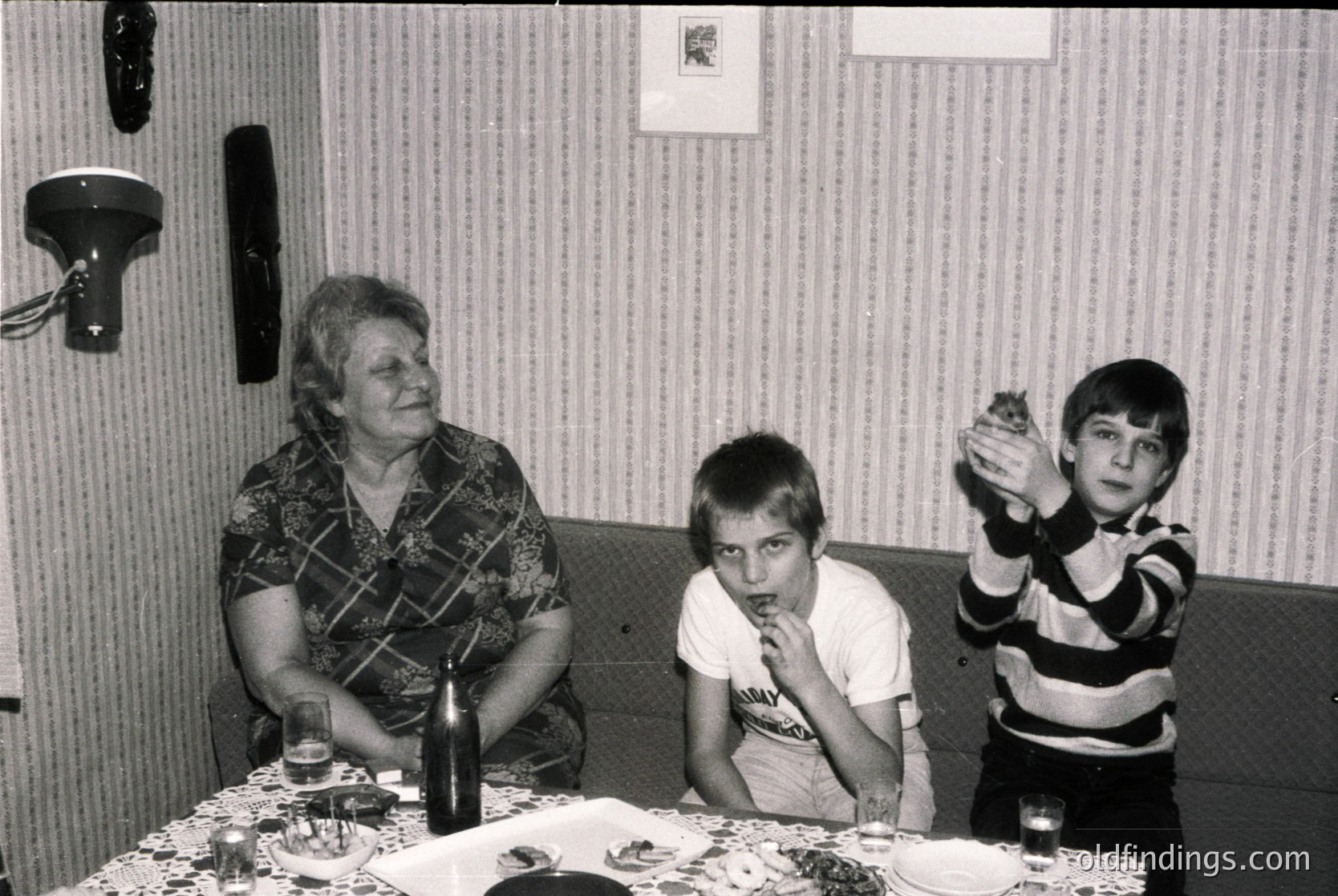 Family gathering in a 1970s-era home, featuring a woman in a patterned blouse and two boys at a table with cake. The striped wallpaper and vintage bottle suggest mid-century domestic life.
