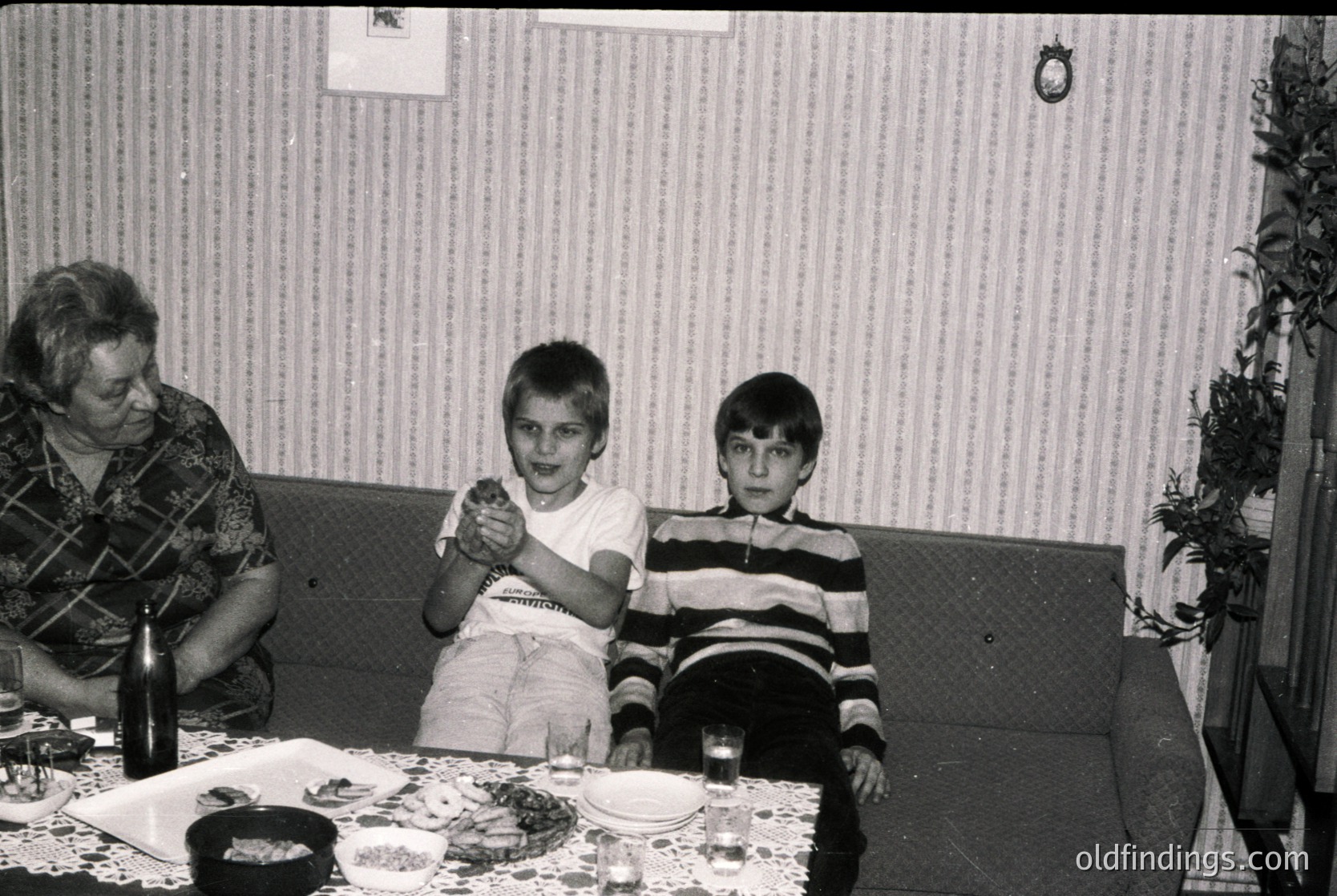 Vintage black-and-white family portrait from the 1970s–80s, featuring a woman and two boys seated at a table with plates of food, glasses, and a bottle. The striped wallpaper and patterned tablecloth suggest mid-century domestic decor. Candid expressions and casual attire evoke nostalgic family moments.