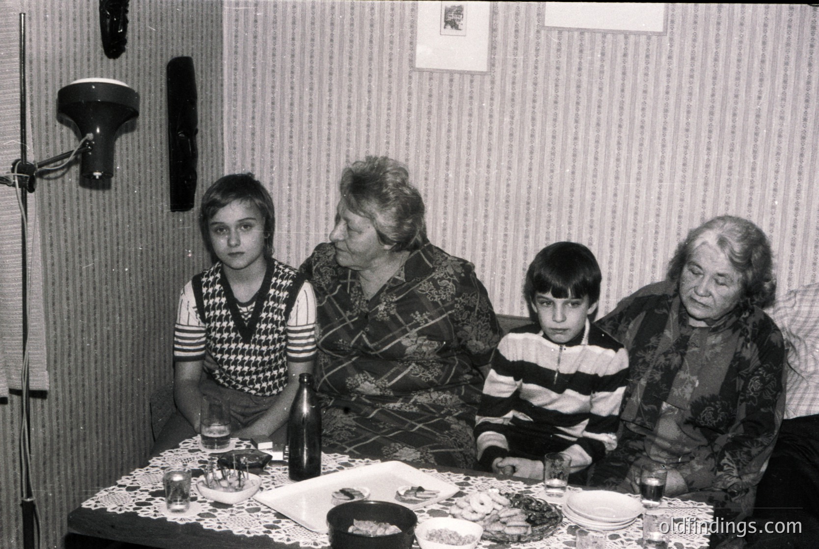 Four generations gathered around a table in a modest indoor setting, likely a 1970s Eastern European home. A woman in patterned sweater and floral blouse sits beside a man in a patterned shirt, flanked by a young girl in striped sweater and a boy in a fur-trimmed coat. Decorative plates, glasses, and a bottle suggest a celebratory meal. Plain wallpaper and a wall-mounted lamp indicate utilitarian decor.