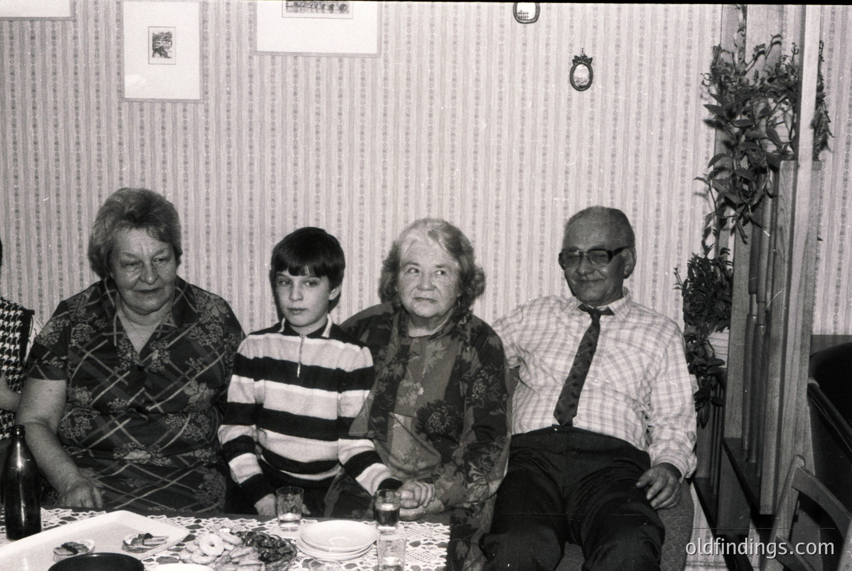 Four generations seated indoors, likely mid-20th century. Elderly woman (left) in patterned sweater, boy in striped shirt, older woman (center) in floral blouse, elderly man (right) in checkered shirt and tie. Plain table with plates, floral wallpaper, and decorative wreath. Warm, intimate family portrait.