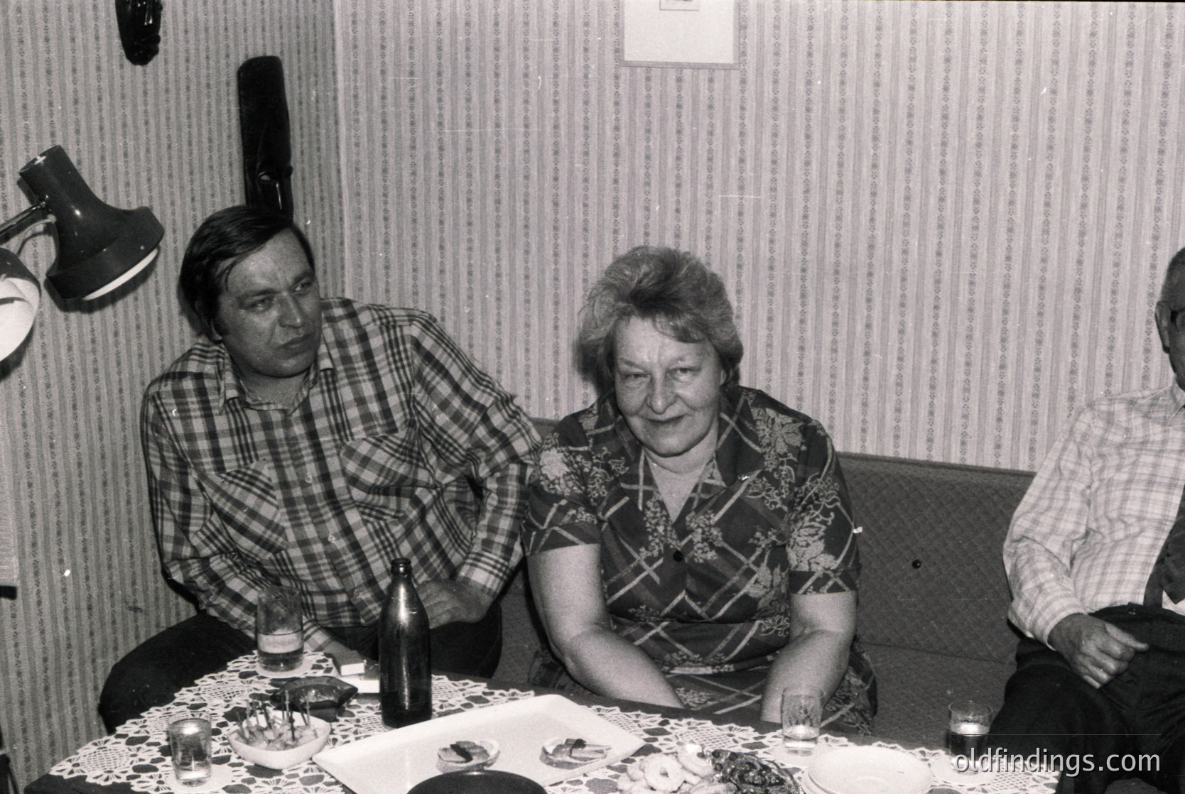 Vintage black-and-white photo of three adults seated indoors, likely mid-1970s Eastern Europe. Woman in patterned blouse examines a jigsaw puzzle; man in plaid shirt leans in, gesturing. Bottles, glasses, and scattered puzzle pieces on table. Strip curtains and desk lamp suggest a domestic or communal setting.