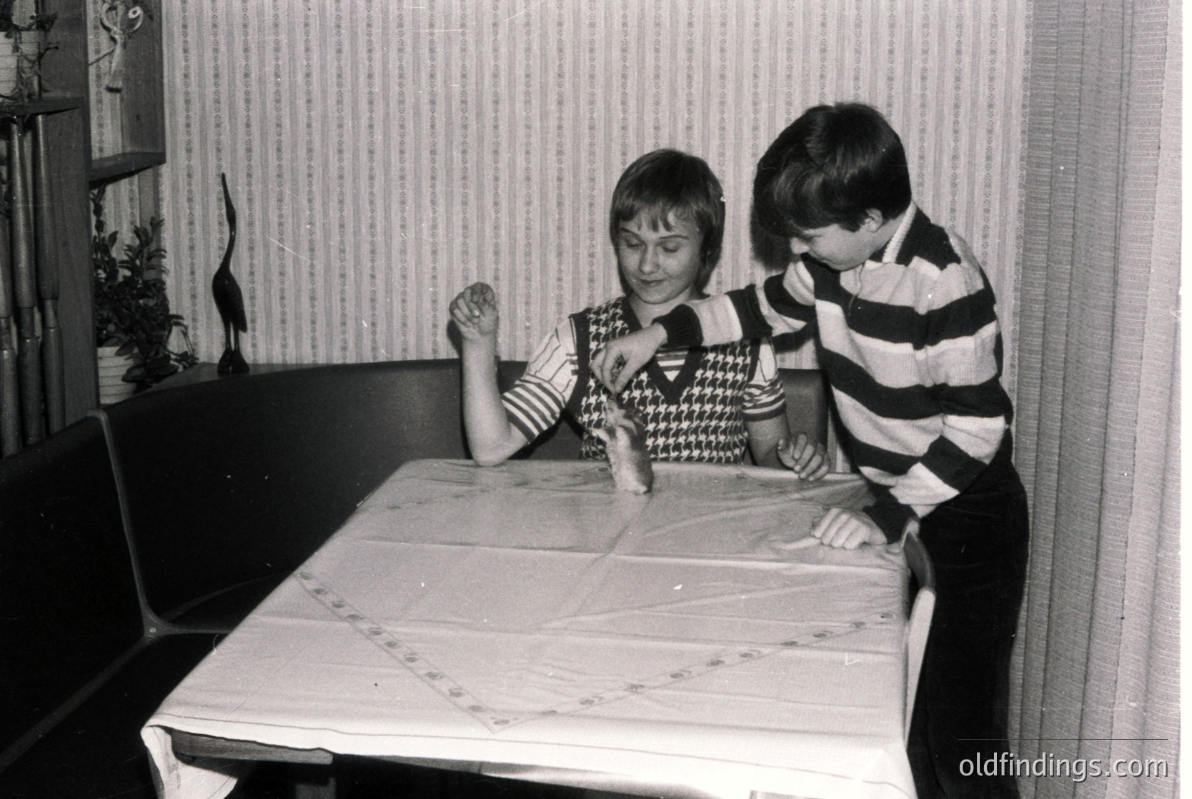 Two children play at a table in a mid-century indoor setting. Girl in patterned sweater leans forward, boy in striped shirt peers intently. Black-and-white photo suggests 1960s–1970s era. Simple wooden furniture and striped wallpaper evoke retro domestic life.
