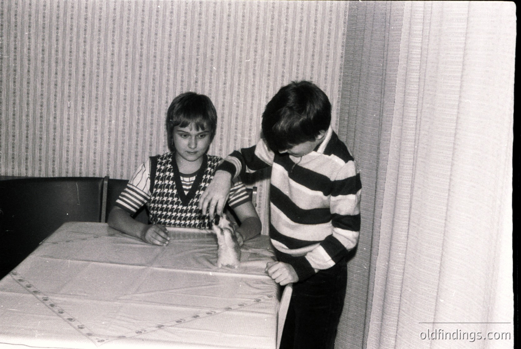 Two boys in 1970s-style striped and patterned sweaters play with a toy train on a tablecloth with floral embroidery. Indoor setting with striped wallpaper. Evokes mid-century childhood nostalgia.