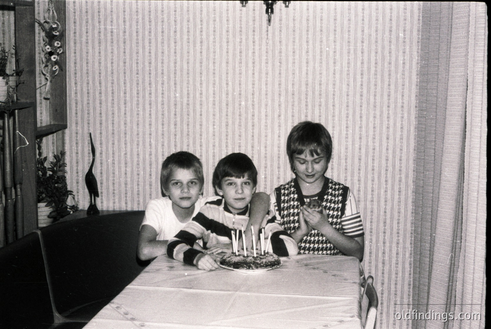 Vintage black-and-white indoor birthday scene: three children pose at a table with a lit cake featuring six candles. Mid-century striped wallpaper and a floral-patterned curtain frame the scene. Simple wooden furniture and a potted plant add context.