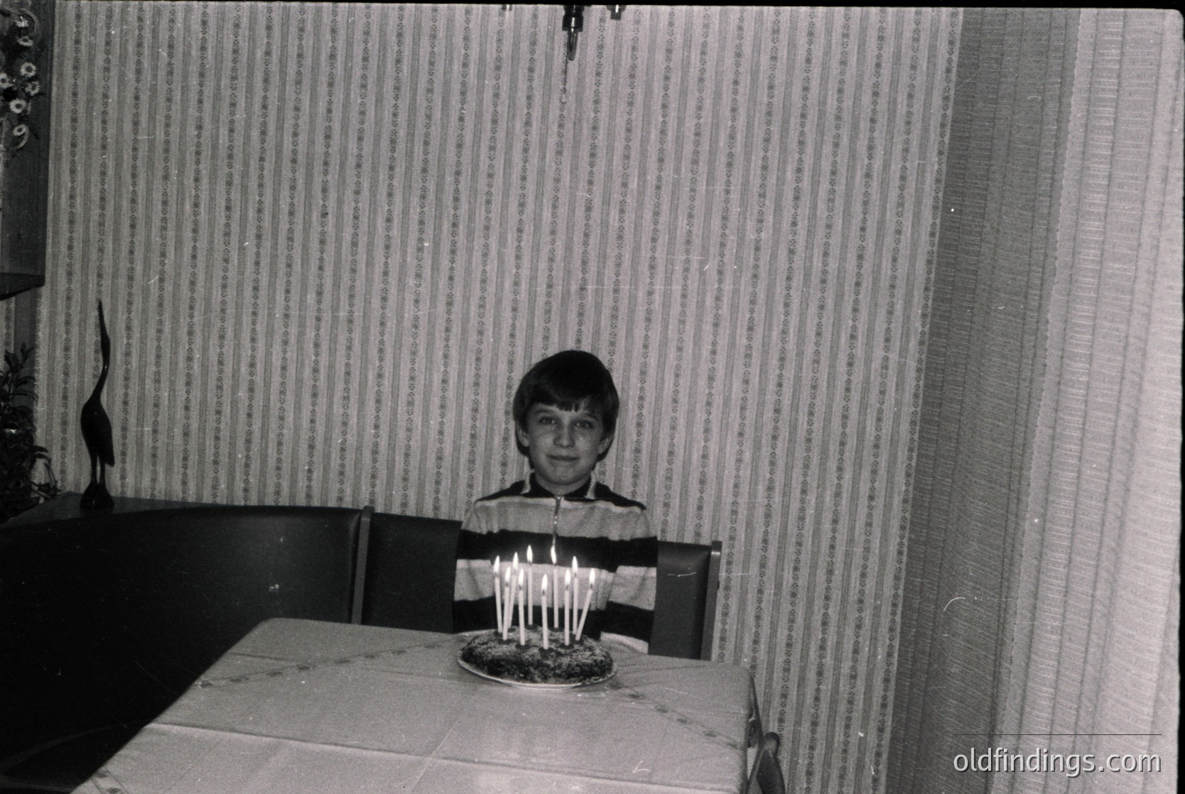 Vintage black-and-white indoor portrait of a young boy seated at a table, blowing out lit candles on a round cake. Plain striped wallpaper and basic wooden furniture suggest mid-20th century domestic setting. Candid, unposed moment captures innocence and nostalgia.