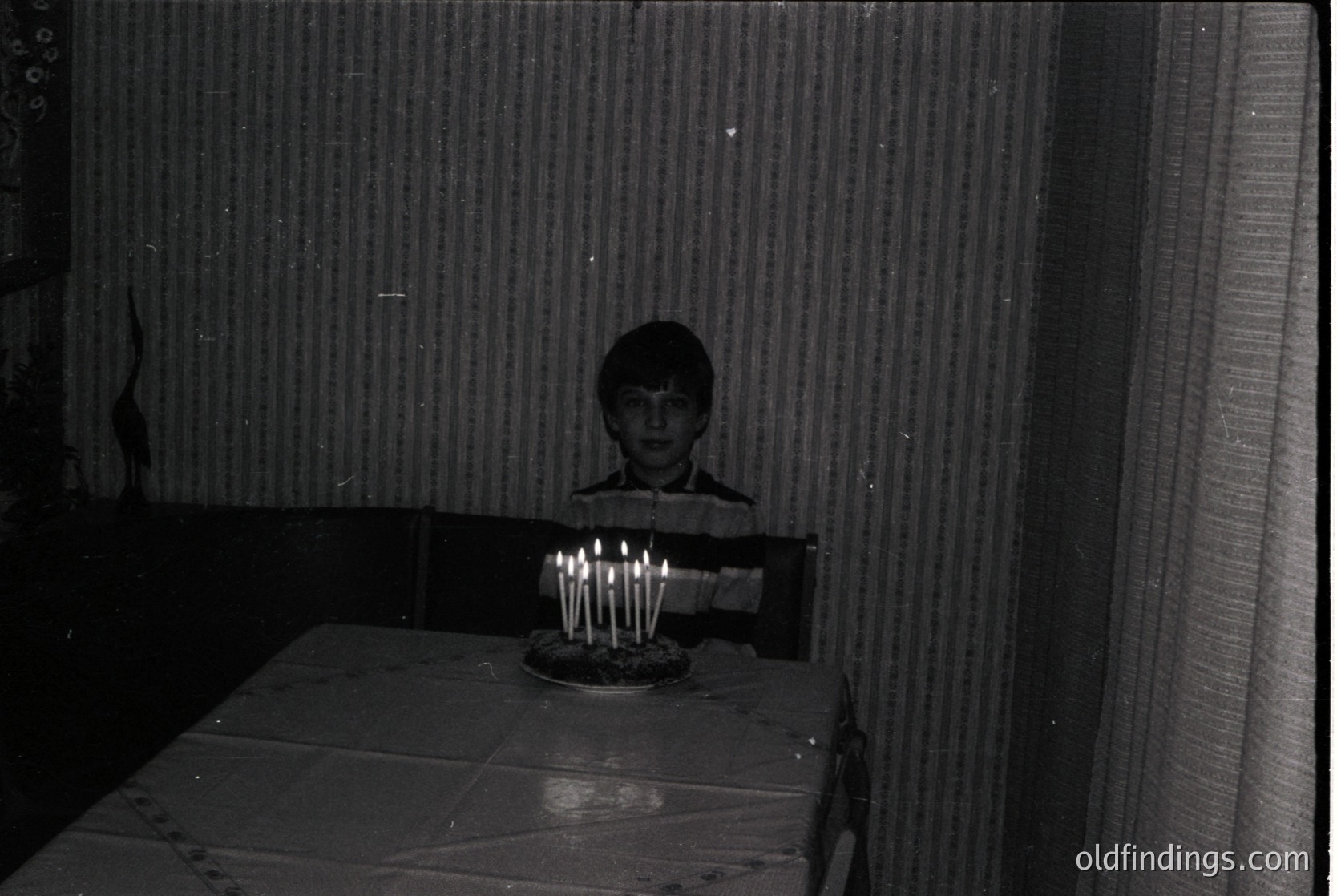Black-and-white indoor portrait of a child standing beside a table with a lit multi-candle birthday cake. Plain wall and curtain backdrop suggests modest 20th-century home decor. Likely mid-century, possibly 1950s–1970s.