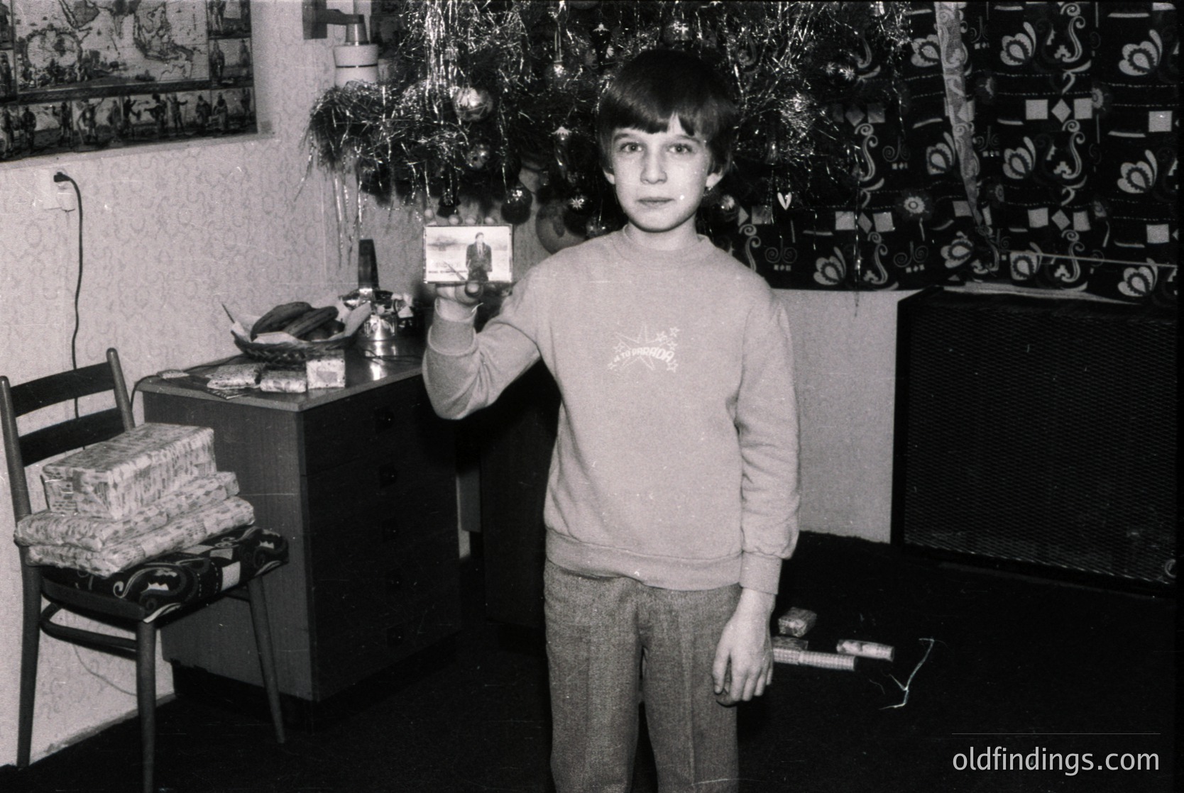 Young boy in 1970s-era sweater holds a small electronic device (likely a calculator or toy) in a modestly decorated indoor setting. Decor includes a Christmas tree, wall art, and a wooden cabinet.